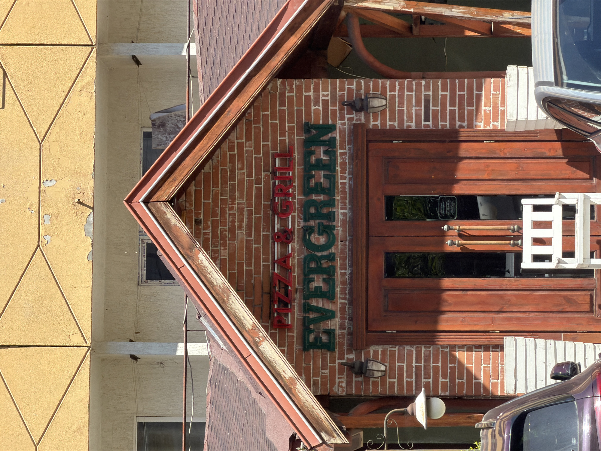 The image captures the entrance of "Evergreen Pizza & Grill" in Sofia, Bulgaria. The building features a brick facade, a wooden-framed entrance with double doors, and signage reading "Pizza & Grill" in red and "Evergreen" in green. The structure has a pitched roof and exterior lighting fixtures. Adjacent to the restaurant, there is a building with a yellow facade. A car is partially visible in the foreground, on the right.