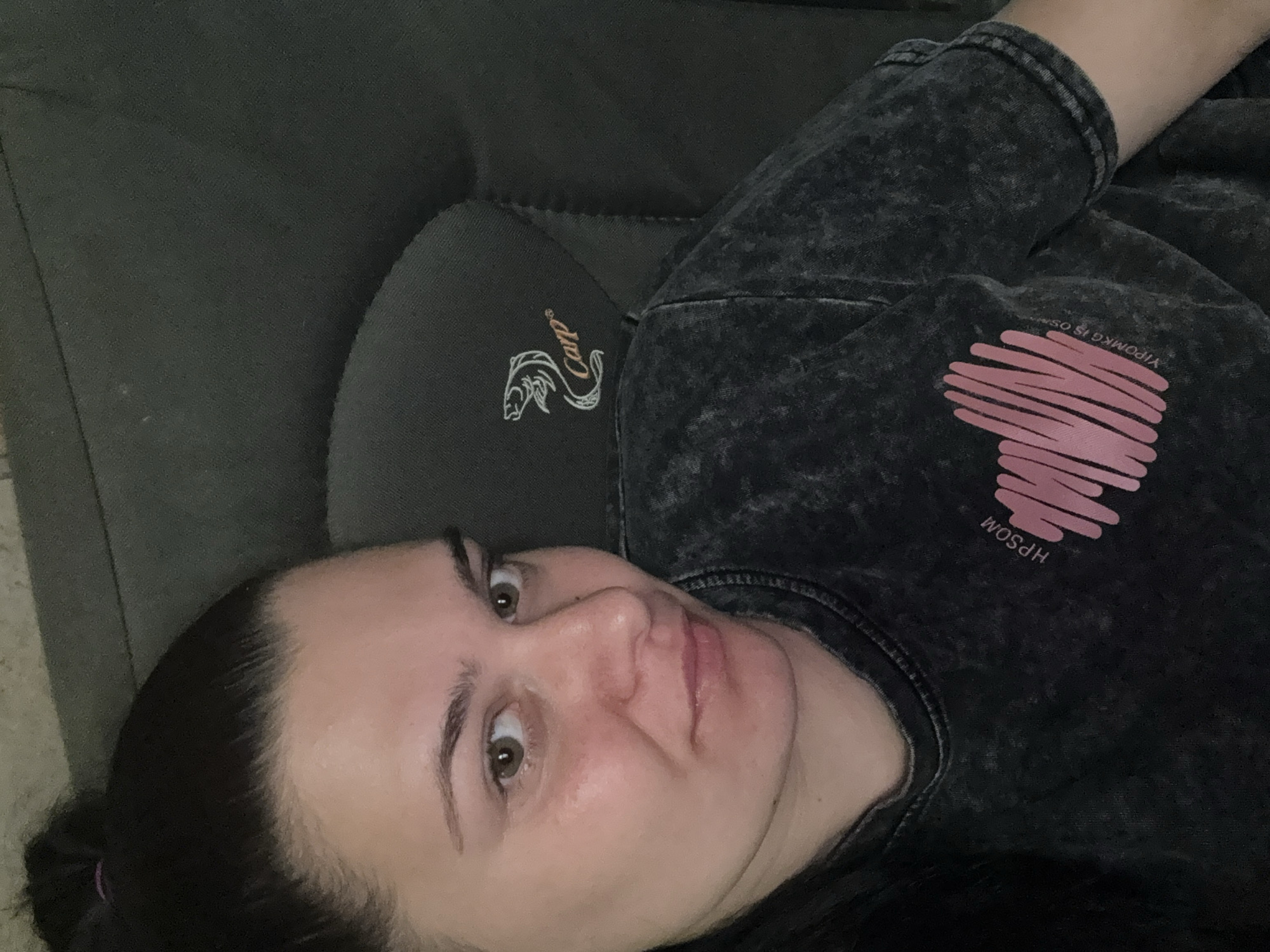 A medium shot captures a woman with dark hair pulled up in a bun and green eyes, looking directly at the camera. She is seated in what appears to be an armchair, indicated by the visible headrest with a "Carp" logo. She is wearing a dark grey, acid-washed t-shirt with a pink heart graphic and text "HPSOM YIPOMRKG IS OS" on the front. The lighting suggests an indoor setting, possibly during daytime. There is no information in the image that indicates the location is the city of Rogosh, Bulgaria.