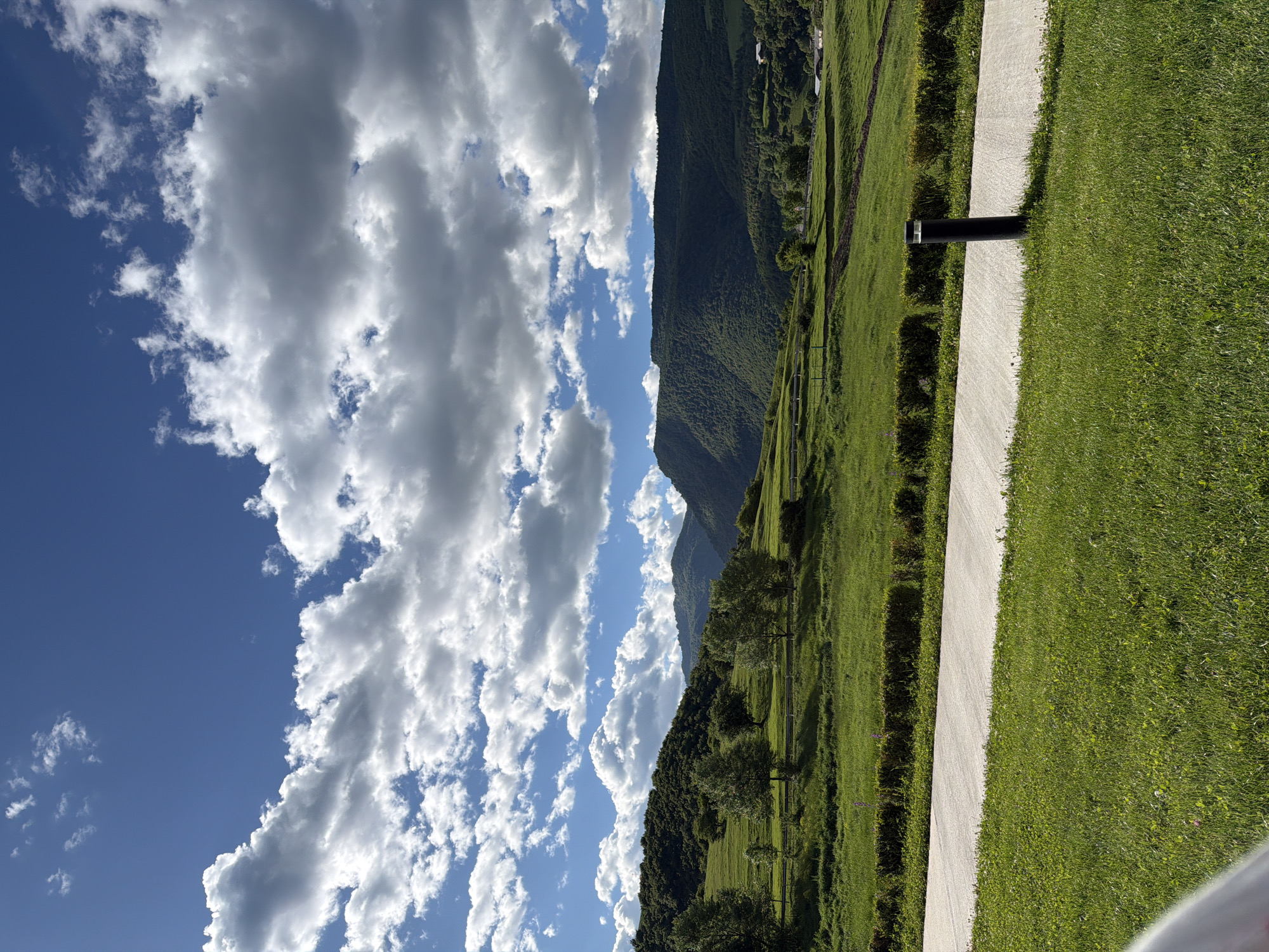 The image depicts a landscape with a clear blue sky adorned with white clouds on the left side, transitioning into a scene of green mountains, grassy fields, and trees on the right. The location is Chkalov, Armenia. A gray pathway cuts through the green grass and a black cylindrical object is positioned beside it. The grassy fields are manicured and bordered by hedges. The mountains are covered in lush green vegetation. The overall atmosphere suggests a serene and picturesque environment.