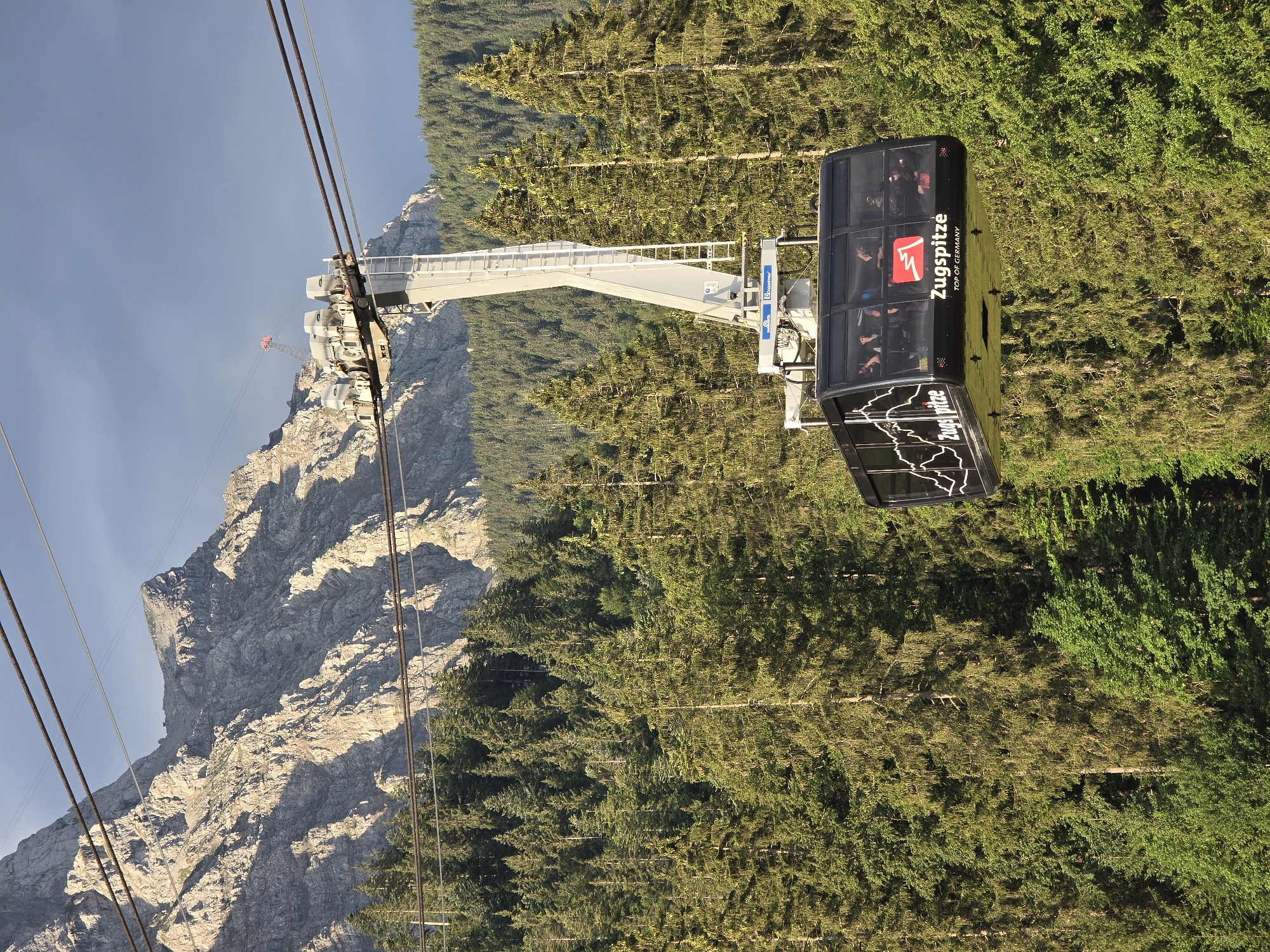 A cable car travels upward in front of a steep, rocky mountain and a dense forest. The cable car is black with large windows, and the word "Zugspitze" is written on its side in white. Inside the cable car, several passengers are visible. The mountain behind the cable car is rugged and illuminated by sunlight, casting shadows. A thick, green forest covers the lower slopes of the mountain and extends into the foreground. The sky is a mix of blue and grey clouds. The scene is likely the Zugspitze cable car in Grainau, Germany, which is a popular tourist destination due to its access to Germany's highest peak.