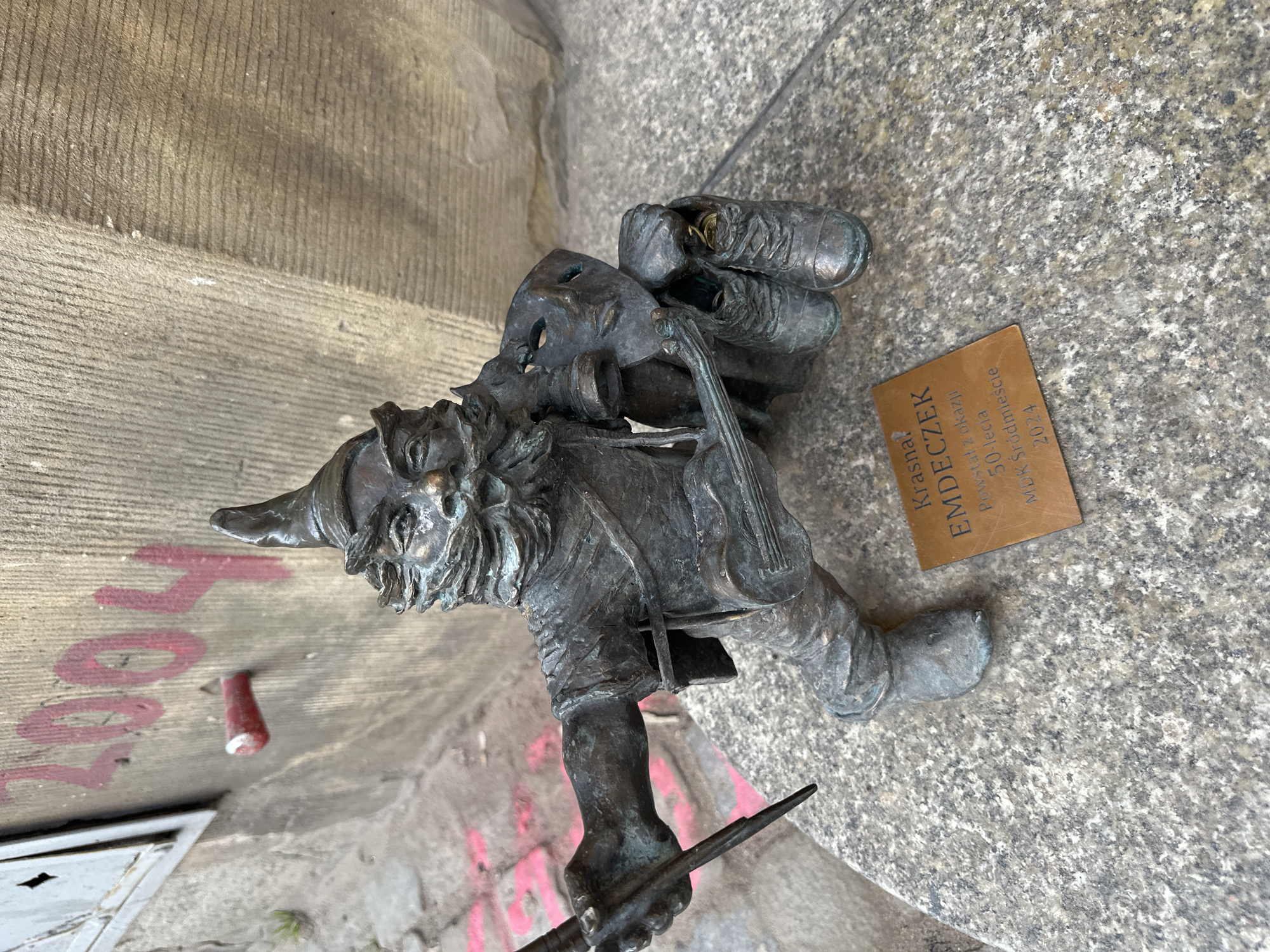 The image shows a bronze statue of a dwarf, one of the many dwarves placed around the city of Wrocław, Poland. This particular dwarf is playing a violin. A plaque nearby reads "Krasnal EMDECZEK Powstał z okazji 50-lecia MDK Śródmieście 2024". The statue is situated on a rough, gray stone surface, possibly a sidewalk or street. The background includes concrete wall elements, some with remnants of graffiti. The statue itself has a theatrical mask and a pair of shoes atop it, indicating a connection to theater. The dwarf is holding a bow in its hand, ready to play the violin.