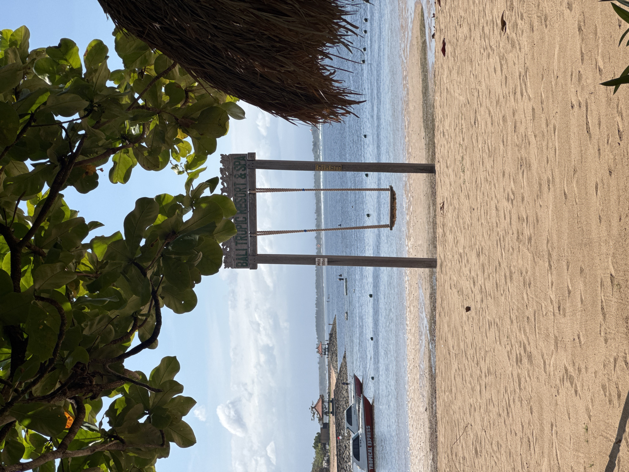 The image depicts a beach scene in Kuta Selatan, Indonesia. The frame is composed of a tree's foliage on the left, a partially visible thatched structure above, and a swing set bearing the words "Bali Tropic Resort & Spa" dominating the central view. Behind the swing set, the ocean meets a cloudy sky. In the lower part of the image, the beach has footprints, and a boat with the words "Tropical Express" is visible. The environment consists of a sandy beach, clear water, and partial views of coastal structures and vessels. There are no people visible.