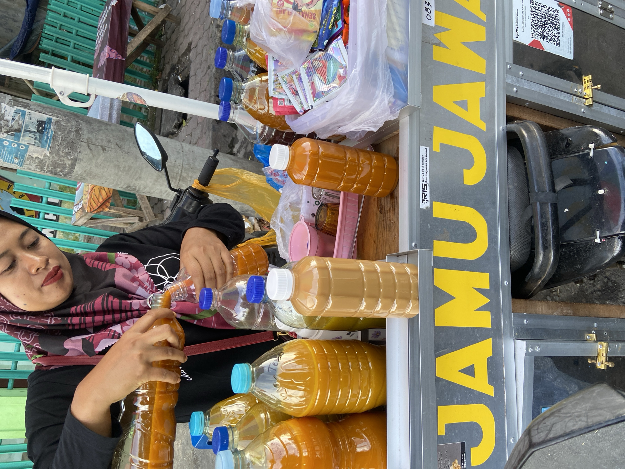 A woman with a red headscarf stands next to a cart of beverages in plastic bottles in Sumbersari, Indonesia. She is pouring a beverage from one bottle to another, presumably for a customer. The cart has several layers with rows of different color beverages. In the background, there are packages of snacks or sweets. The side of the cart has the words "JAMU JAWA" in large yellow letters.