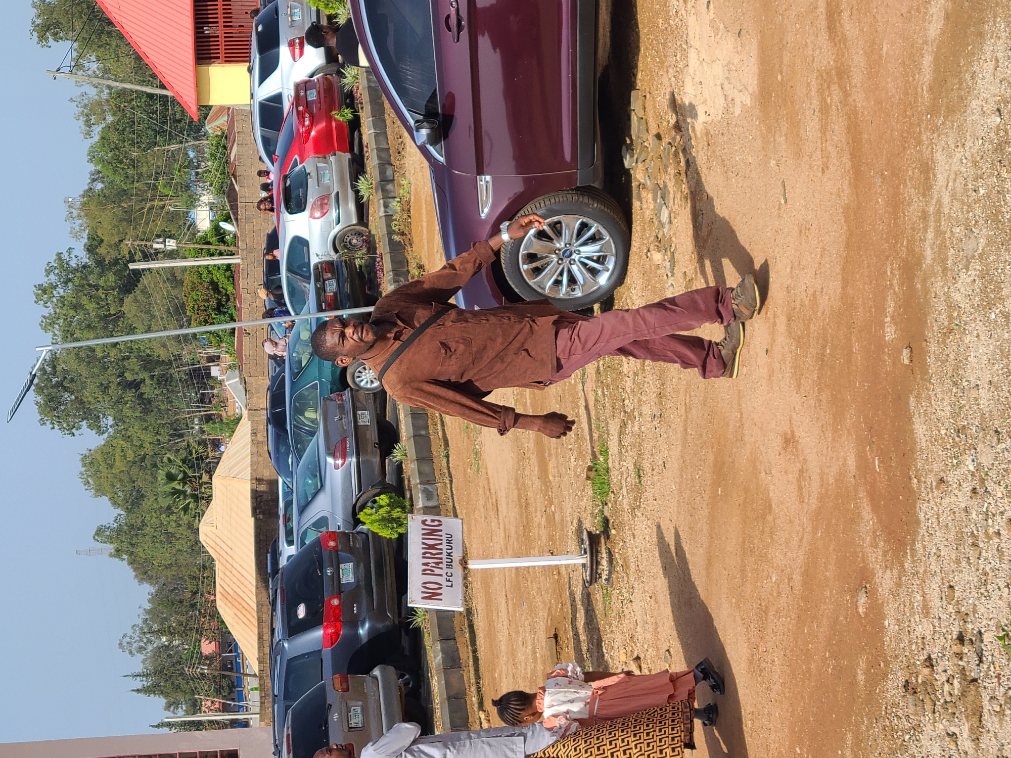 A group of people are gathered in a dirt parking lot in Gura Top, Nigeria, with numerous cars parked in the background. A man in a brown shirt and maroon pants walks towards the foreground, his left hand resting on the hood of a maroon car. A young girl in a pink and white dress stands to his left, facing away from the camera. A "NO PARKING LFC BUKURU" sign is visible on a white pole in the middle of the lot. Buildings and trees form the backdrop under a clear sky.