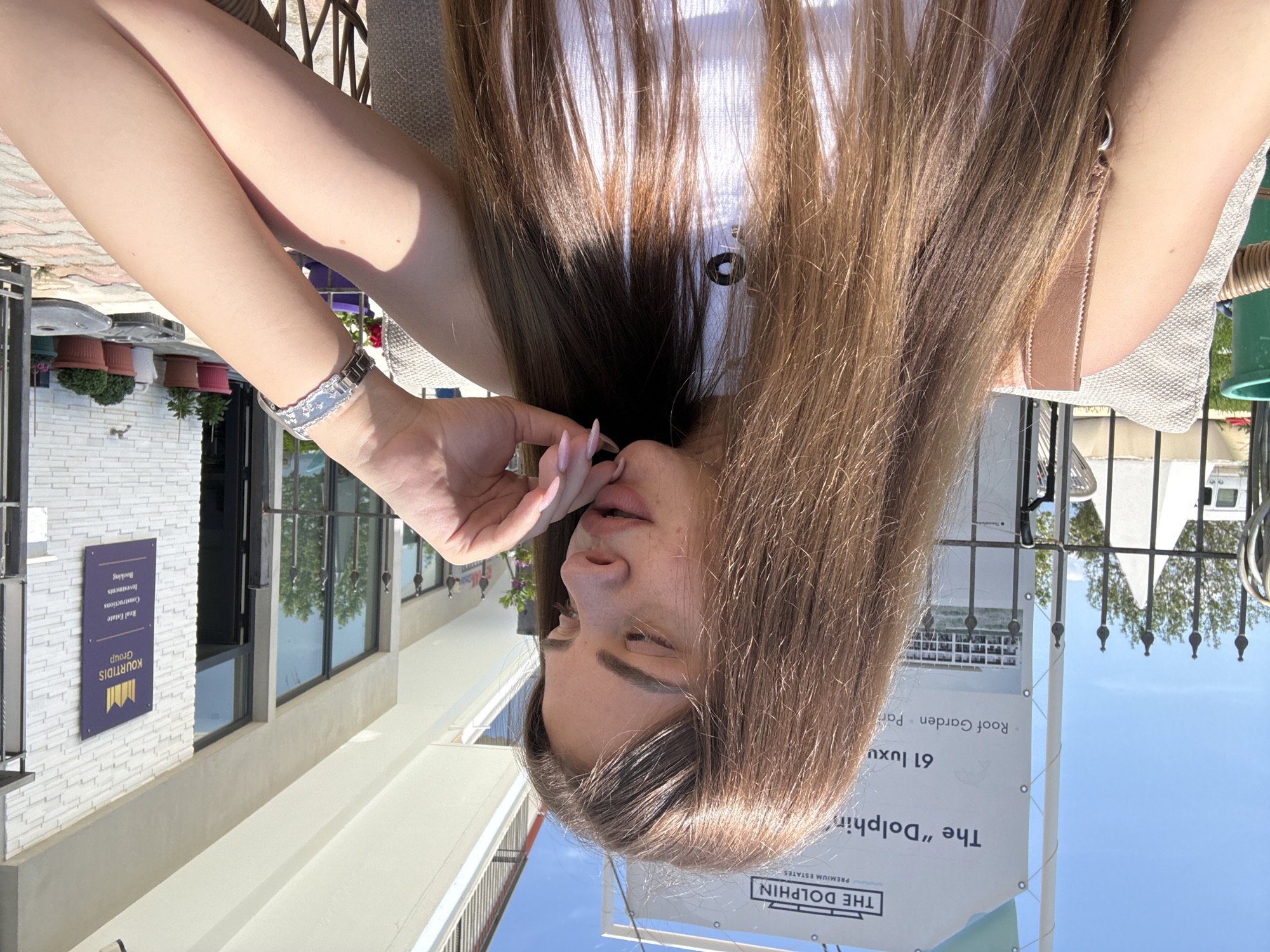 Here is a description of the image:
The image shows a woman with long brown hair sitting outside. The location appears to be an urban area with buildings and outdoor advertising present. One building has a sign identifying the "Kourtidis Group" involved in real estate, construction, investments, and booking. A nearby sign advertises "The Dolphin" premium estates with "61 luxu roof garden par." The woman is seated, wearing a white top, and has her hand near her face. The overall setting appears to be a relaxed outdoor area in a city.
