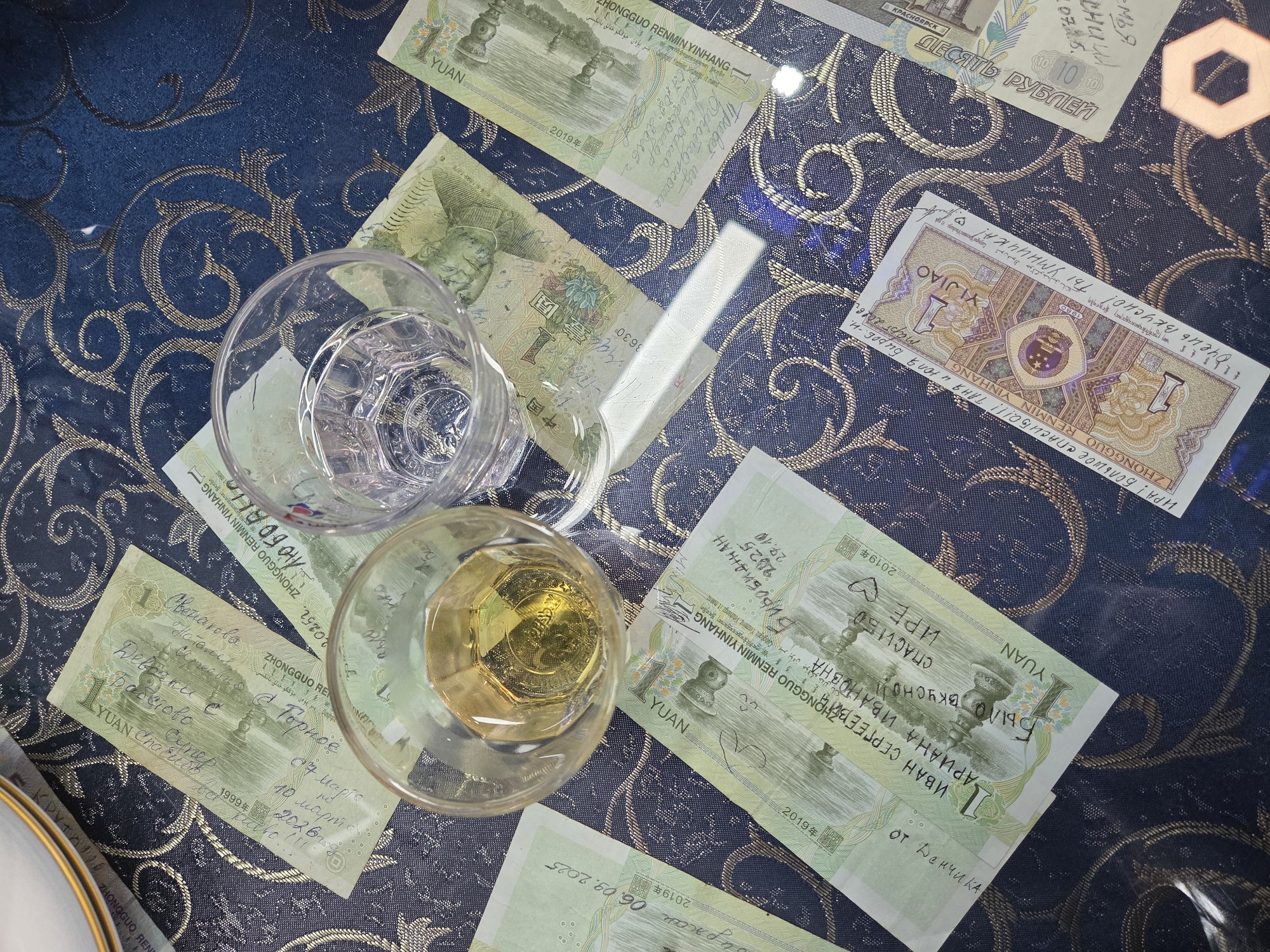 The image displays a collection of banknotes, predominantly Chinese Yuan and one Russian Ruble, scattered across a dark blue table with an intricate gold and bronze patterned design. Two glasses are also on the table; one is clear and empty, while the other holds an amber-colored liquid.
The scene appears to be set indoors, likely at a dining table, suggested by the tablecloth and glasses. No people are visible, but the handwritten notes on the banknotes indicate human interaction and appreciation. The mix of Chinese and Russian currency, along with extensive Russian handwritten text, strongly suggests a location where both cultures and languages intersect, such as Tongjiang, China, a city bordering Russia.
Notable details include:
* **Banknotes:** Several 1 Yuan Chinese banknotes (from years 1999 and 2019) are visible, along with one 1 Jiao Chinese banknote and one 10 Russian Ruble banknote (featuring Krasnoyarsk). Many banknotes have handwritten messages.
* **Glasses:** One clear, empty glass with a hexagonal base pattern, and another glass containing a golden-brown beverage, possibly an alcoholic drink, reflecting light from its hexagonal base.
* **Handwritten text:** Numerous messages in Russian are scrawled across the banknotes. These include expressions of gratitude like "СПАСИБО" (Thanks), "Было вкусно" (It was tasty), and "Очень вкусно! Ты УМНИЧКА!" (Very tasty! You're a smart girl!). Names such as "ИРА," "Таня и Юля," "Наталья Сергеевна," "ИВАН СЕРГЕЕВИЧ," "АРИАНА ИВАНОВНА," and "От Денчика" are present. Dates like "10 марта 2026," "06.09.2025," "27.10 2025," and "07.25." are also written, along with location references like "Биробиджан" and "Привет из Биробиджана" (Greetings from Birobidzhan).
* **Currency text:** Chinese characters (e.g., 壹圆, 中国人民银行), and text in Uyghur, Tibetan, Mongolian, and Zhuang scripts are visible on the Yuan notes, alongside "YUAN" in Latin script. The Ruble note clearly states "ДЕСЯТЬ РУБЛЕЙ" (Ten Rubles).
The overall impression is that of gratuities or personal notes left after a meal or gathering, reflecting appreciation and personal connections between individuals, possibly in a cross-cultural context.