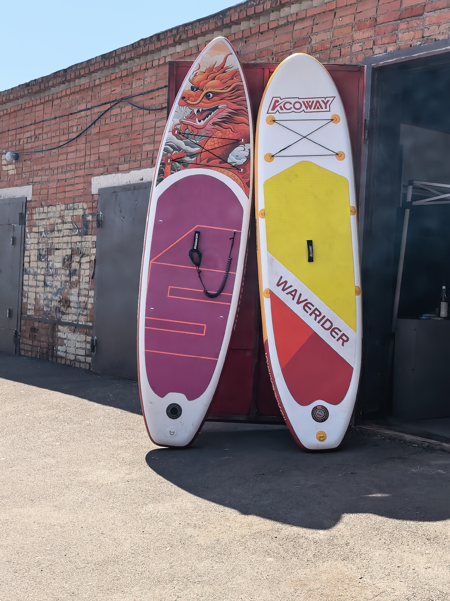 Two stand-up paddleboards are leaning against a red brick wall next to an open garage door. The board on the left has a purple section with pink lines and a large red dragon graphic at the top. The board on the right is white with a yellow section at the top and a red and white design at the bottom, featuring the text "WAVERIDER." Both boards have black bungee cords on their decks. The ground is paved with asphalt. The scene appears to be outdoors in a sunny urban environment.