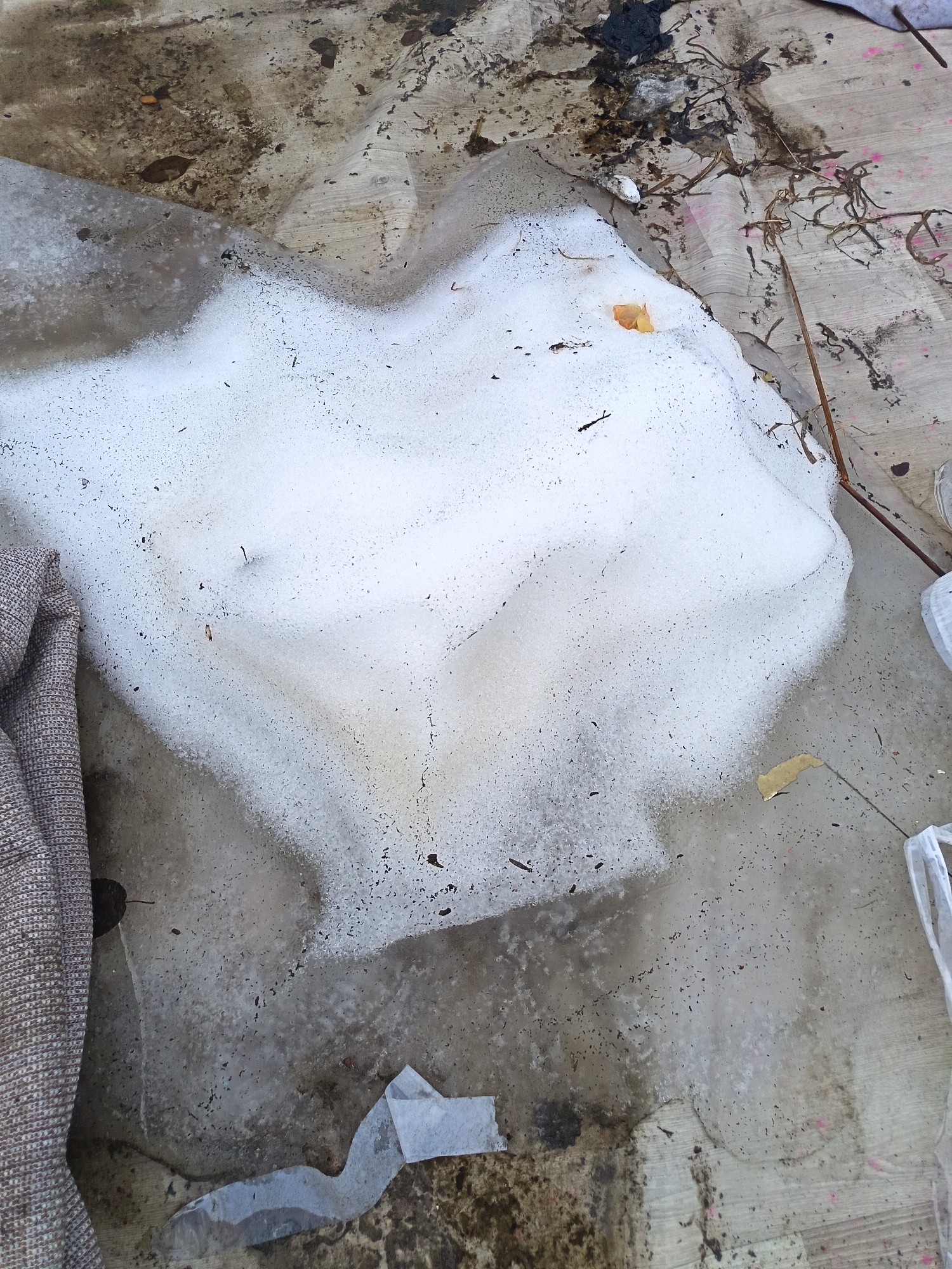 The image depicts a patch of melting snow on a dirty surface, likely an exterior floor or ground area. The snow is irregularly shaped, with areas of full coverage and areas where it has melted away, revealing the underlying surface. The surface appears to be concrete or a similar material, stained and marked with dirt, debris, and possibly some discoloration. A piece of fabric or cloth is visible on the left edge of the frame. There are various small pieces of debris scattered around, including twigs and small scraps of paper or plastic. The lighting suggests an overcast day or a location in shade. The composition and subject matter suggest a state of neglect or disuse. There is no information available to corroborate a location in Nizhnevartovsk, Russia.