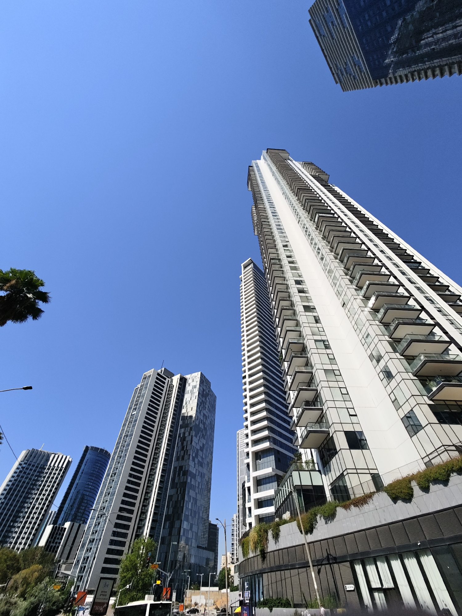 The image shows a low-angle view of modern skyscrapers in Givatayim, Israel, under a clear blue sky. The buildings are predominantly white and glass, with many balconies. In the foreground, there are trees and some greenery. The image captures a street-level perspective with traffic lights, street lamps, and what appears to be a bus. One building in the upper right corner is darker and more reflective, contrasting with the lighter buildings. The overall scene depicts a contemporary urban environment.