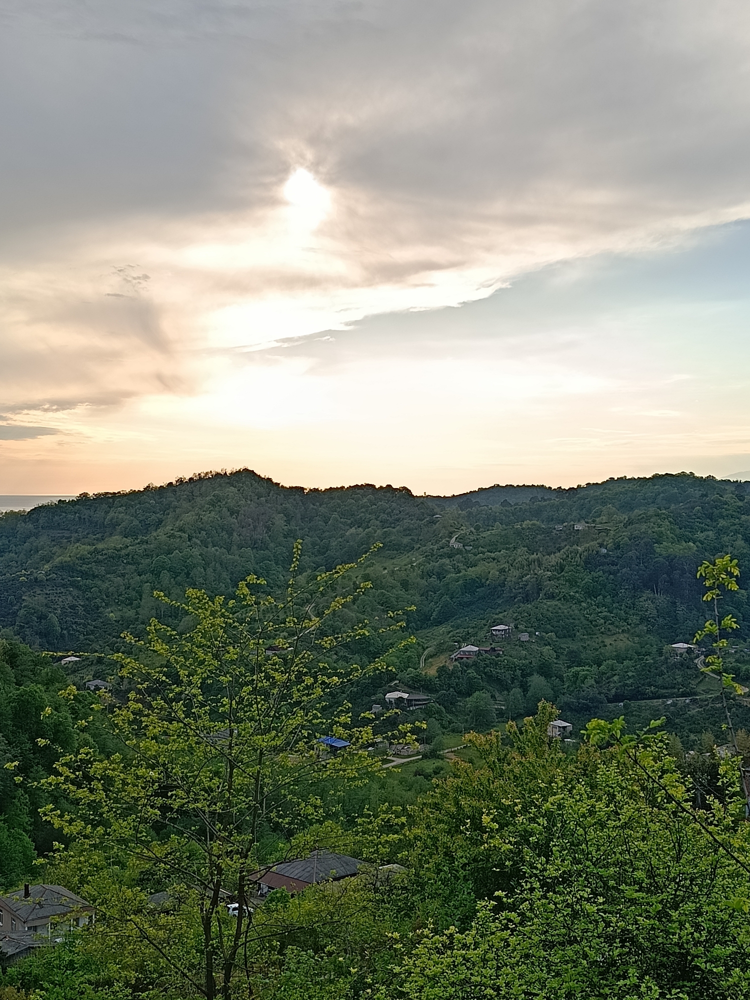 The image shows a hilly landscape in the city of Yuada-Gulrypsh, Abkhazia. Lush green vegetation covers the hills, with trees dominating the landscape. Several houses are scattered throughout the area. The sky is overcast with a break where the sun is visible.
