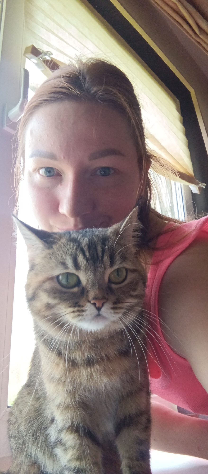 A woman is holding a tabby cat in front of her. The woman has fair skin and blue eyes, and her reddish-brown hair is pulled back. She is wearing a pink tank top. The cat has green eyes and tabby markings. They are near a window. The location is Unknown, Unknown.