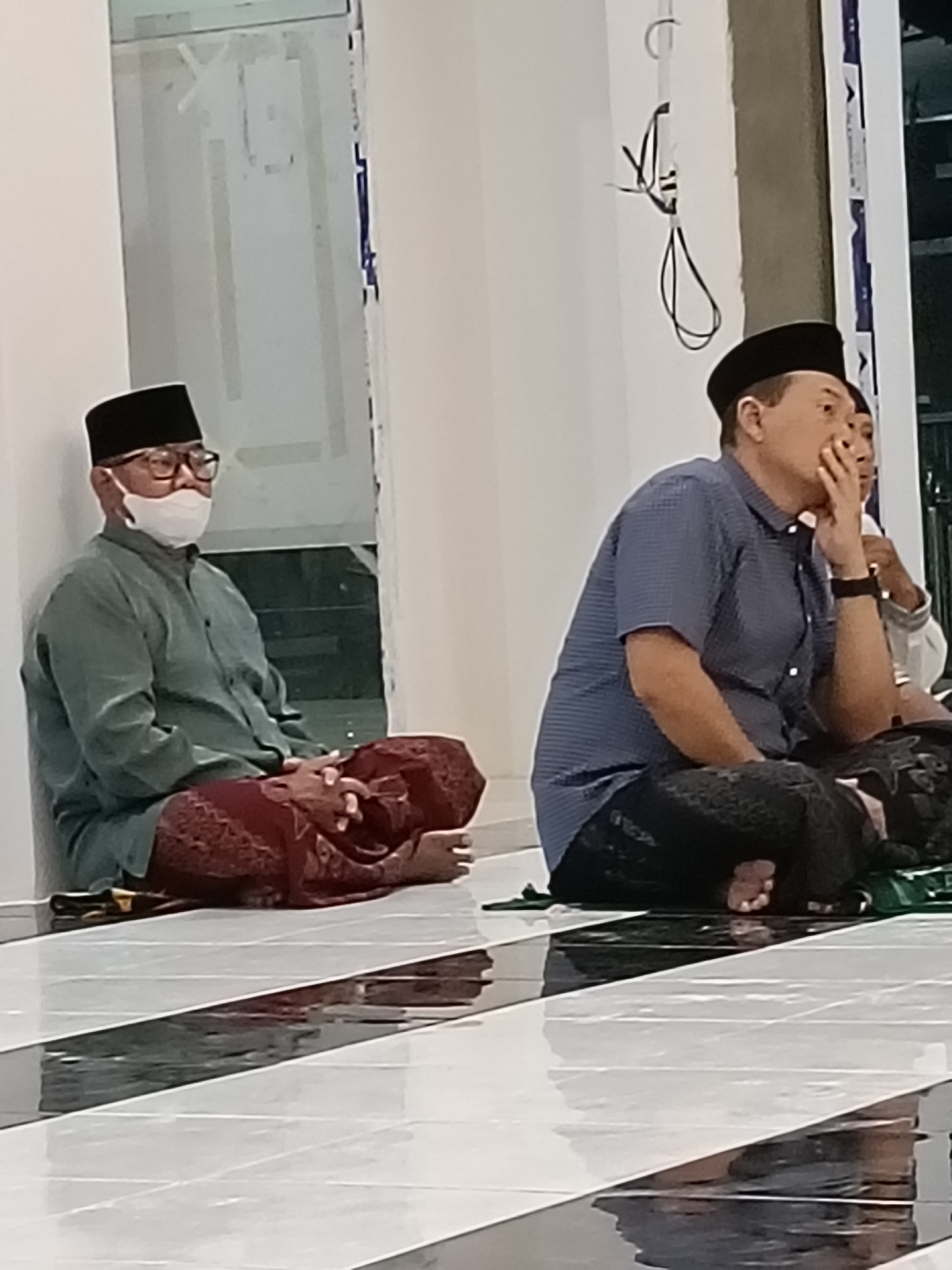 Two men are sitting on the floor in a building, possibly a mosque, in Kediri City, Indonesia. The man on the left is wearing a green shirt, a red sarong, a black peci (cap), and glasses, with a white mask covering his nose and mouth. He is leaning against a white wall with his legs crossed and his hands resting on his lap. The man on the right is wearing a blue checkered shirt, black pants, and a black peci. He is resting his chin on his hand, looking to his right. The floor is tiled with black and white diagonal stripes, and the tiles reflect the overhead lighting. The background shows a wall and some electrical wiring hanging from the ceiling.