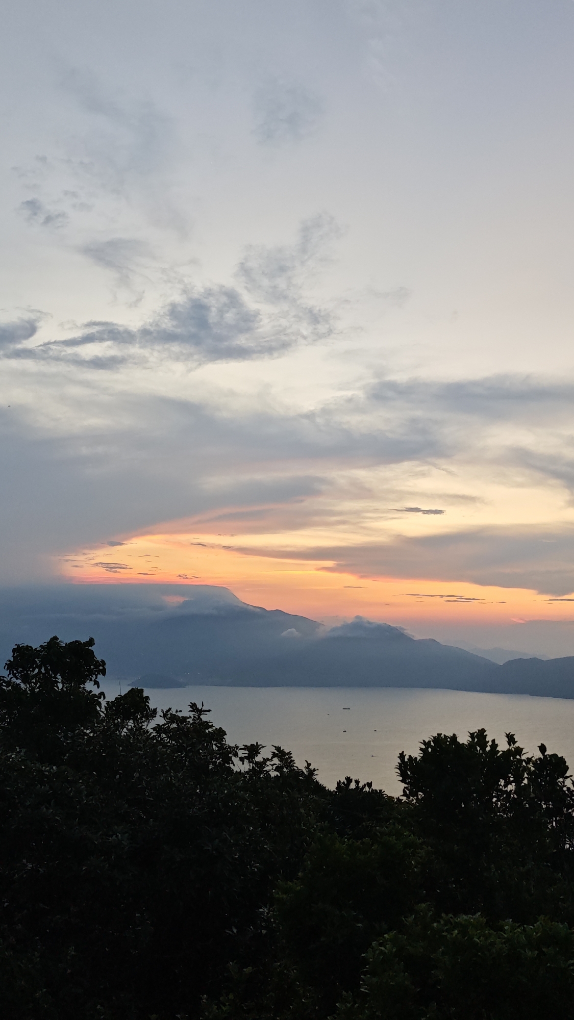 The image shows a landscape view at sunset or sunrise, with dark green foliage in the foreground, likely from trees or shrubs. The middle ground features a large body of water, possibly a bay or lake, with a mountain range in the distance. The mountains appear partially obscured by clouds or fog. Above the mountains, the sky transitions from pale yellow and orange near the horizon to shades of gray and blue higher up, indicating the time of day. There are a few small vessels visible on the water. This is an elevated view likely from a mountain top around the city of Đà Nẵng, Vietnam.