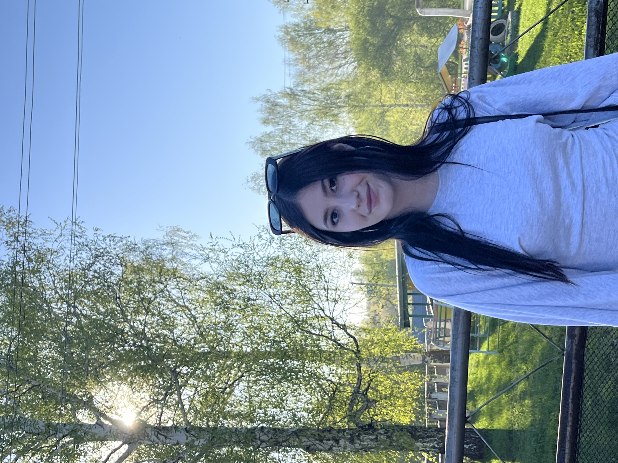 A young woman with long, dark hair is posing outdoors. She is wearing sunglasses on her head and a long-sleeved light grey shirt. The background includes a tree with the sun shining through its leaves, a green grassy area, a metal fence, and power lines overhead. The location is in Фоки, Russia.