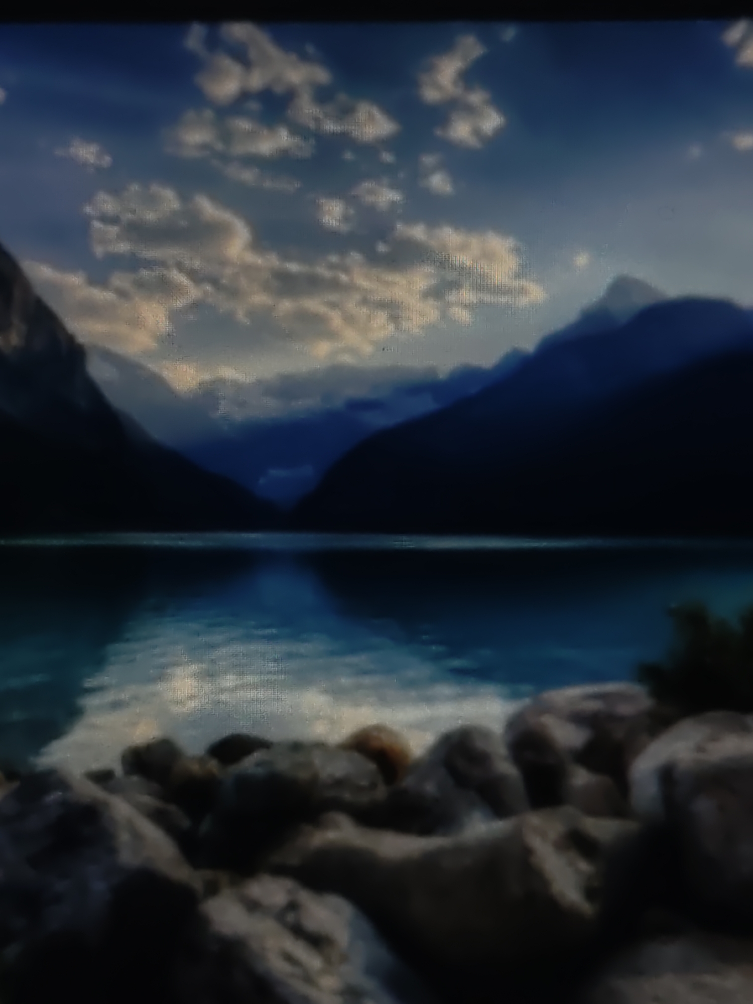 The image depicts a serene natural landscape. In the foreground, there is a rocky shore with numerous boulders of varying sizes and textures. Beyond the shore lies a calm body of water, reflecting the sky and the surrounding mountains. The water appears deep blue and still, with subtle ripples breaking the perfect reflection. In the background, imposing mountains rise, their dark, silhouetted forms contrasting with the bright, cloudy sky. The sky is a mix of deep blue and white, with fluffy clouds scattered across it, illuminated by sunlight. The overall scene evokes a sense of tranquility and natural beauty. The location is not specified in the city of Kurganinsk, Russia, as the image portrays a wilderness landscape.