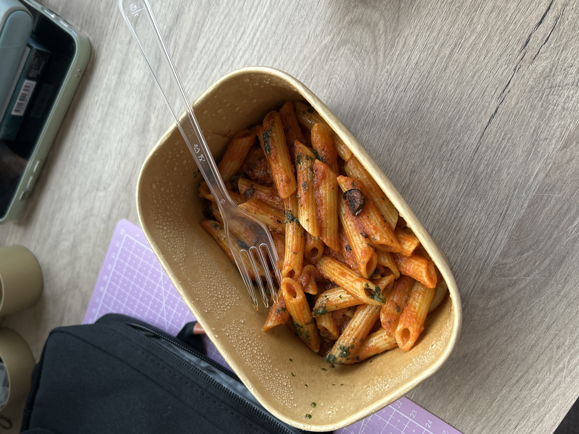 A top-down view depicts a container of penne pasta with tomato sauce and green herbs on a table, with a clear plastic fork inserted into the pasta. The container is rectangular with rounded corners and a light brown color, showing condensation droplets on the exterior. The pasta is orange-red in color from the sauce.
To the left of the pasta, there's a smartphone with a light green case, a beige cylindrical object (possibly a container or part of a larger object), and a black zippered bag. Beneath the container and bag is a purple cutting mat with visible measurement markings (22-25). The tabletop has a light brown wood-grain pattern.
The image does not allow for determining the location in Sofia, Bulgaria. No people, interactions, or specific environmental details beyond the immediate tabletop are visible.
