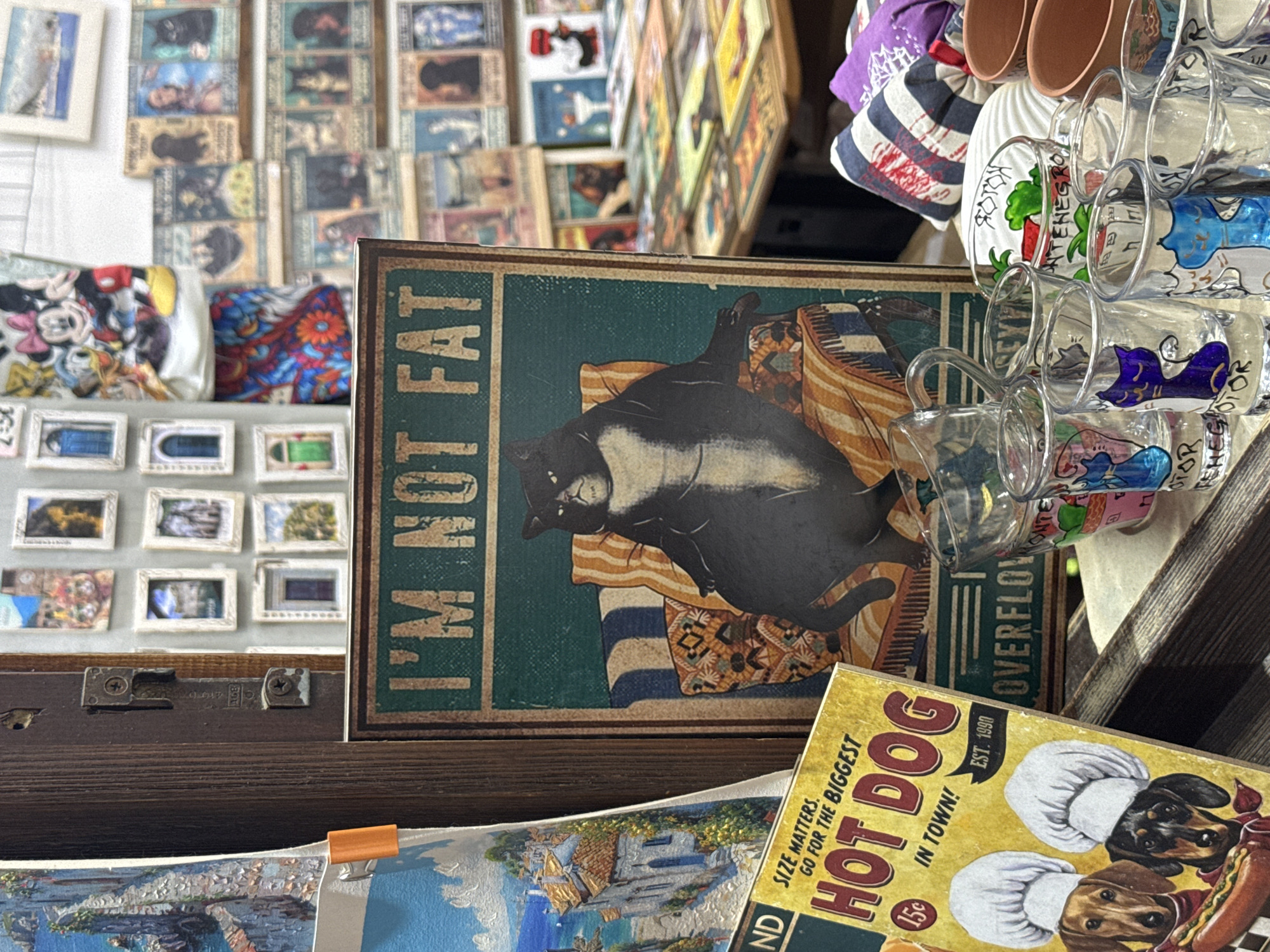 The image displays a close-up view of a market stall in Kotor, Montenegro. The foreground is dominated by a framed sign featuring a cartoon of a fat black cat sitting on a striped couch, with the text "I'M NOT FAT" prominently displayed above it. To the left of this sign is a postcard or small poster depicting a picturesque coastal town with white buildings and blue accents, possibly a view of Kotor itself, with the text "SIZE MATTERS. GO FOR THE BIGGEST HOT DOG IN TOWN!" and "EST. 1990".
Further to the right and slightly behind, a collection of small glass shot glasses are arranged. Several of these glasses have hand-painted designs of cats in various colors and text that appears to include "MONTENEGRO". Some of the glasses also have images of food items like strawberries. In the background, a wall is covered with small framed pictures or postcards, many of which appear to be of scenic locations or possibly vintage advertisements. A table laden with more items, including what looks like more postcards and some pottery, is visible in the far background. The overall impression is of a souvenir stall offering a variety of quirky and themed items. No people are clearly visible in the image.