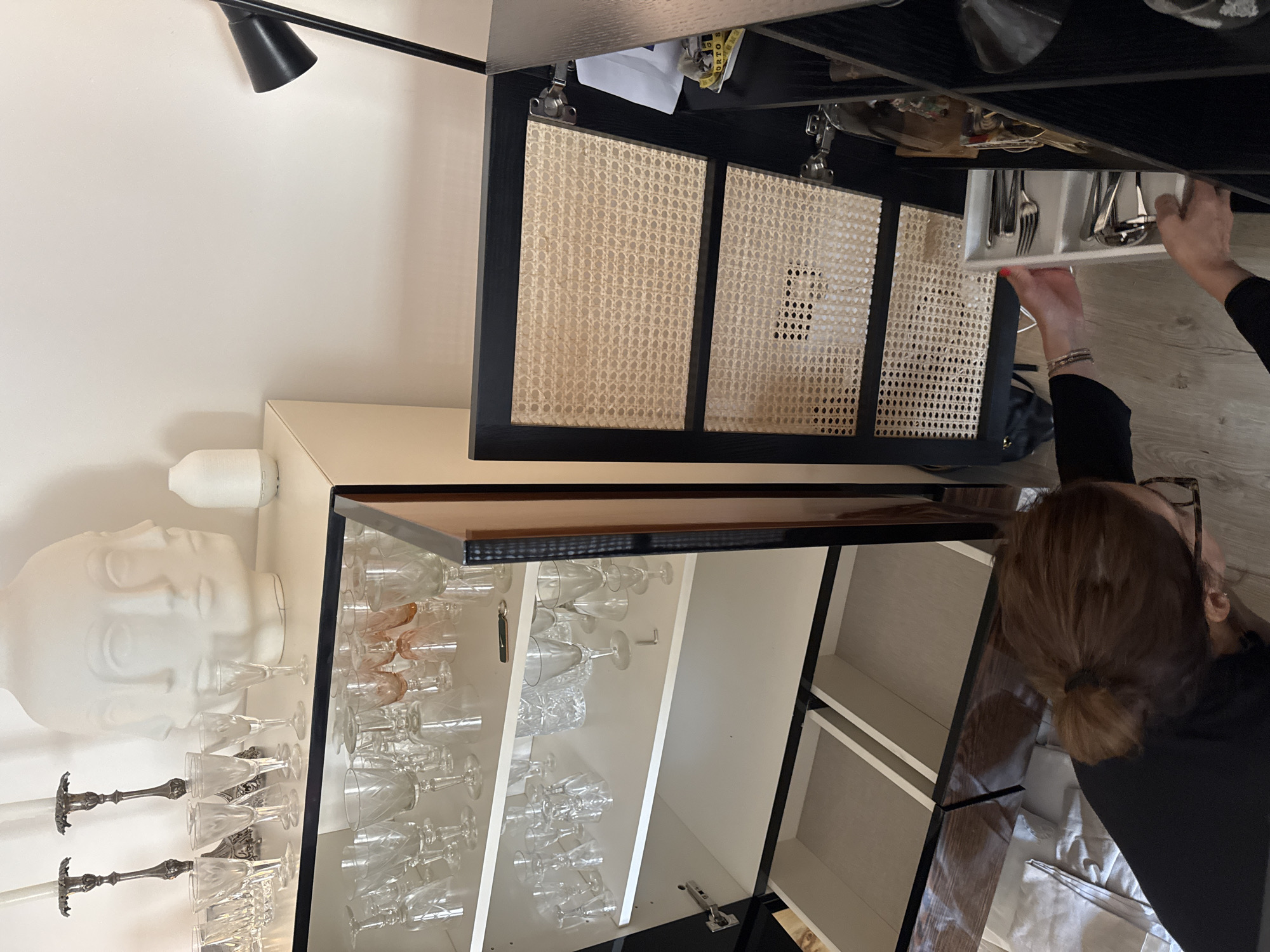 The image shows a room interior, seemingly in a residential setting, located in Vila Nova de Gaia, Portugal. A person is visible, with brown hair pulled back in a low bun, wearing glasses and a black shirt. The individual is interacting with a cabinet that is partially open. The cabinet is black and white, with a top portion containing a drawer with woven panels and a lower portion displaying numerous wine glasses and other glassware on shelves. A white bust, a pair of candelabras, and a white lamp are situated on the cabinet's side or above it. The cabinet appears to be well-stocked and maintained. A black desk lamp is also visible near the top of the frame. The person appears to be taking out silverware from a drawer.