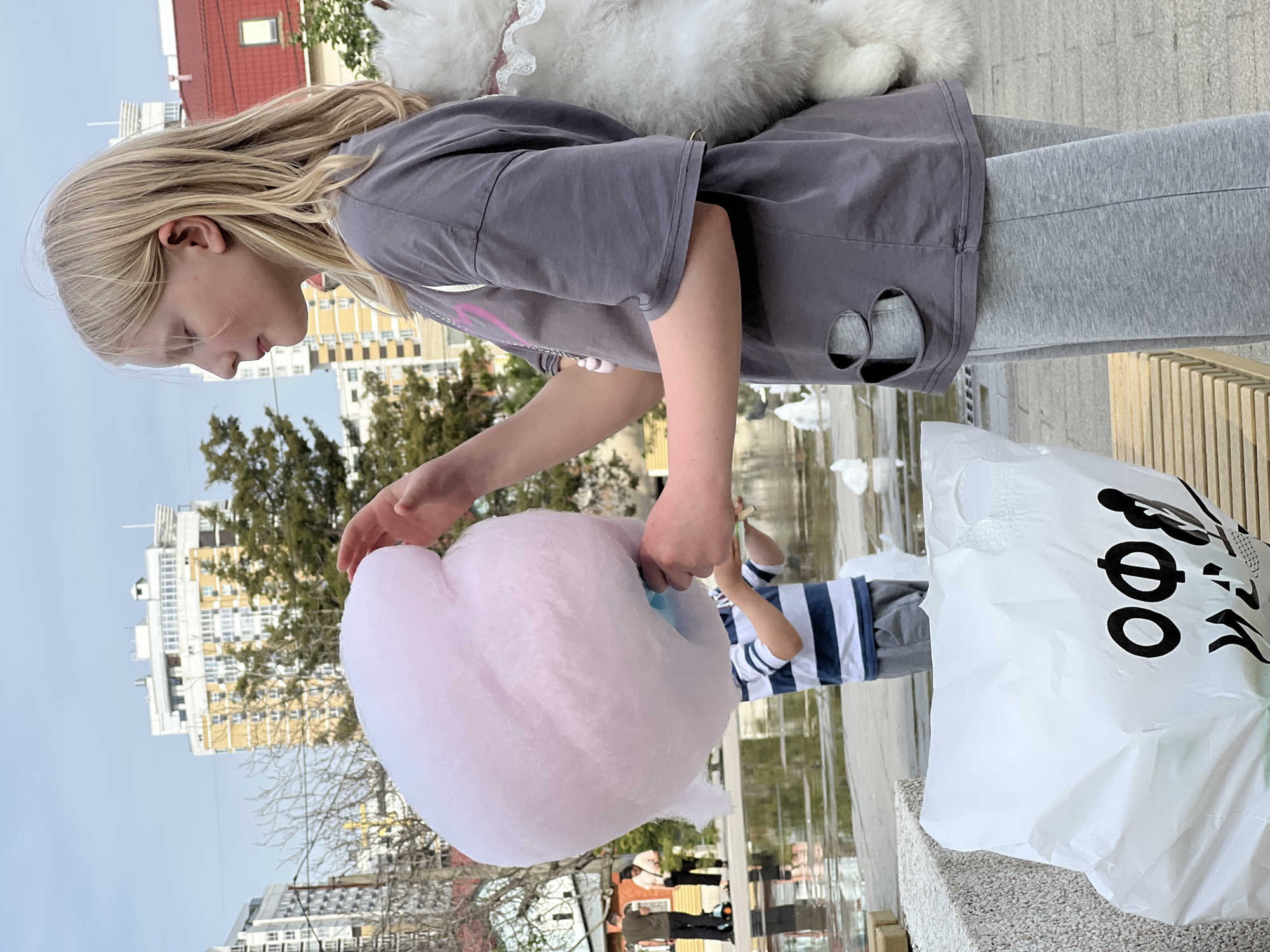 A young girl with blonde hair is holding a large pink cotton candy in her right hand. She is wearing a grey t-shirt and light grey sweatpants. Behind her is a white fluffy object, possibly a toy or an article of clothing. To the right, a white plastic bag with black writing on it rests on a light-colored surface. Another child in a blue and white striped shirt stands behind the girl. In the background, buildings are visible, suggesting an urban setting. Bare trees and a clear sky indicate it might be spring or fall. The scene takes place outdoors. The image is rotated. The buildings in the background suggest this might be in Krasnodar, Russia.