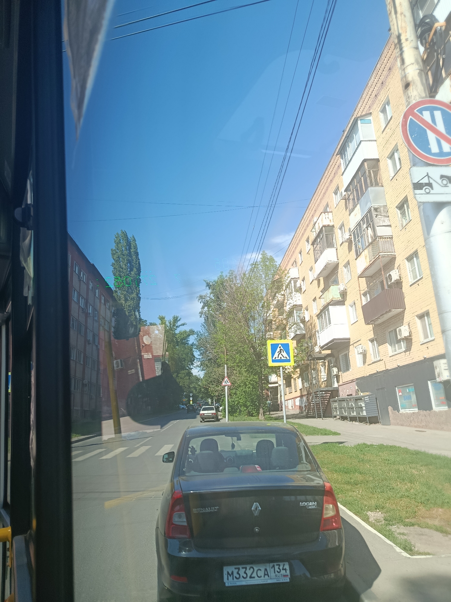 The image depicts a street scene in Saratov, Russia, viewed from inside a vehicle, likely a bus or a car, through a window that reflects the sky and surrounding buildings. A dark-colored sedan, identified by the license plate "M332CA 134 RUS", is in the foreground, stopped at a crosswalk. The crosswalk is clearly marked with white stripes on the asphalt. On the right side of the street, a tall, yellow brick apartment building with multiple balconies is visible. Several air conditioning units are mounted on its exterior. A traffic sign with a red circle and a diagonal red bar over a blue background with white vertical lines indicates a prohibition, and below it, a sign depicts a tow truck, suggesting a no-towing zone. Further down the street, another apartment building, appearing older and brick-faced, is on the left. Trees and greenery are present along the sidewalks and between the buildings. The sky is clear and blue. The overall impression is a typical urban environment on a sunny day.