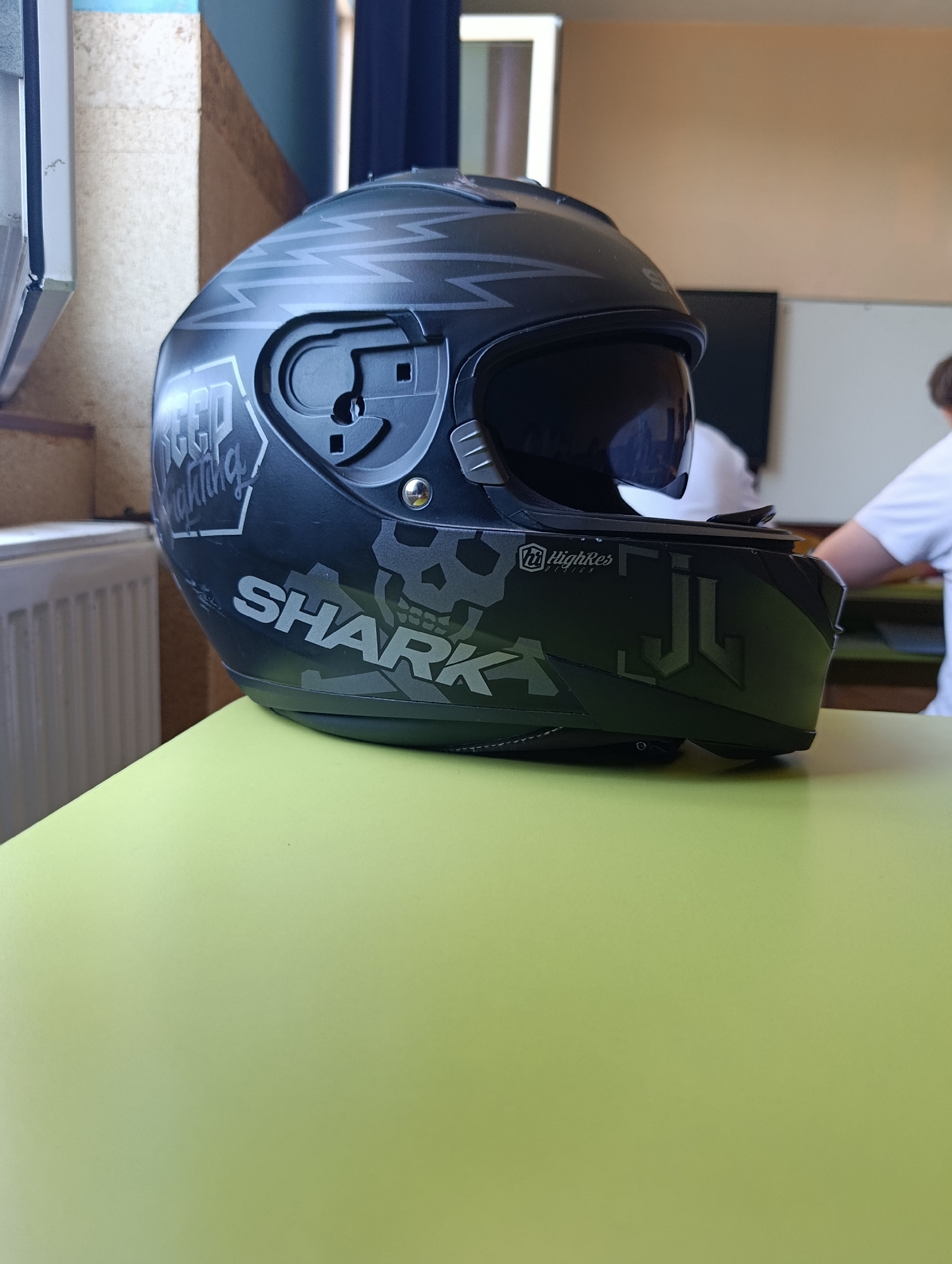 A black motorcycle helmet with a dark visor is placed on a light green surface. The helmet has a complex grey graphic design, including lightning bolts and a skull motif. The brand "SHARK" is visible in large grey lettering on the side of the helmet, along with smaller logos, one of which reads "HighRes". To the left of the helmet, a white radiator is visible, and to the right, blurred figures of people wearing white shirts are present in what appears to be an indoor setting. The background consists of a wall with a diagonally patterned section and a dark blue curtain. The scene is indoors, possibly a classroom or a common area, and is not specifically indicative of Stamboliyski, Bulgaria, beyond being a generic interior space.