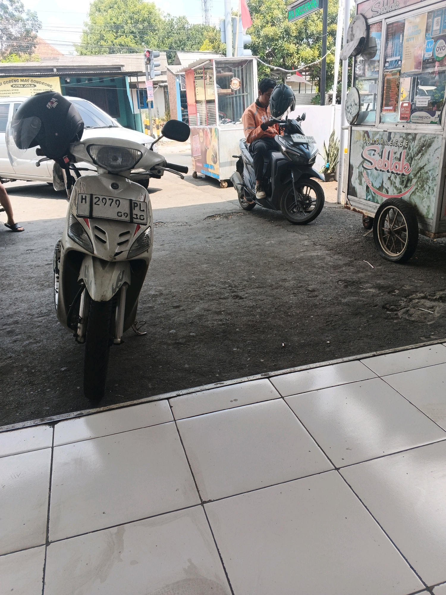 In the city of Genuk Barat, Indonesia, the image depicts a street scene dominated by two parked motorcycles. In the foreground, a white scooter with license plate H 2979 PL is prominently displayed, its front end angled slightly towards the viewer. A black helmet rests on the handlebars of this scooter. Behind it, a dark-colored motorcycle, likely a sport bike, is occupied by two individuals. The rider, wearing an orange jacket and dark pants, is seated on the bike, holding a black helmet. The second person, also with a helmet, is seated behind the rider.
To the right of the motorcycles, a food cart with "SUNDANESE SEBLAK" branding is parked. The cart has a large, white body with artistic green and white graphics and is equipped with two wheels. The background reveals a typical street environment with buildings, trees, and a traffic light, indicating an urban setting. The ground surface is a mix of asphalt and a tiled area in the immediate foreground. The lighting suggests it is daytime, with sunlight casting shadows.