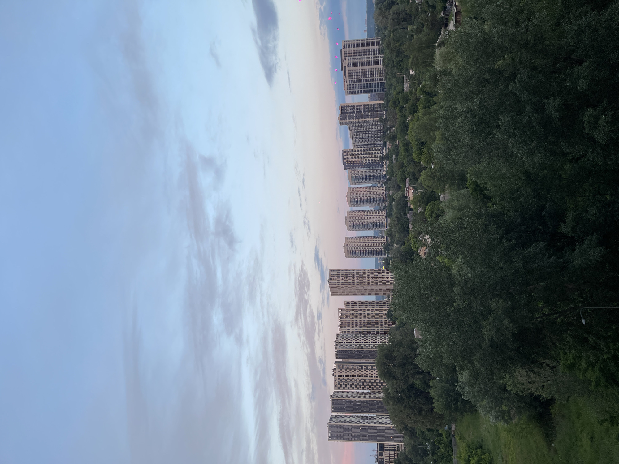This is an outdoor, aerial, full shot of a city skyline in Kyiv, Ukraine, during dusk. The sky is a gradient of pale blue to pale pink, with scattered, wispy clouds. In the middle ground, a cluster of modern, high-rise apartment buildings with patterned facades stands out against a backdrop of dense green trees. The buildings vary in height and architectural design, with some featuring intricate window arrangements. In the foreground, the tops of large, dark green trees are visible, obscuring the lower parts of the cityscape and indicating a position elevated above the immediate surroundings. The overall atmosphere is serene and urban, with the natural elements of trees and sky contrasting with the constructed environment of the city.