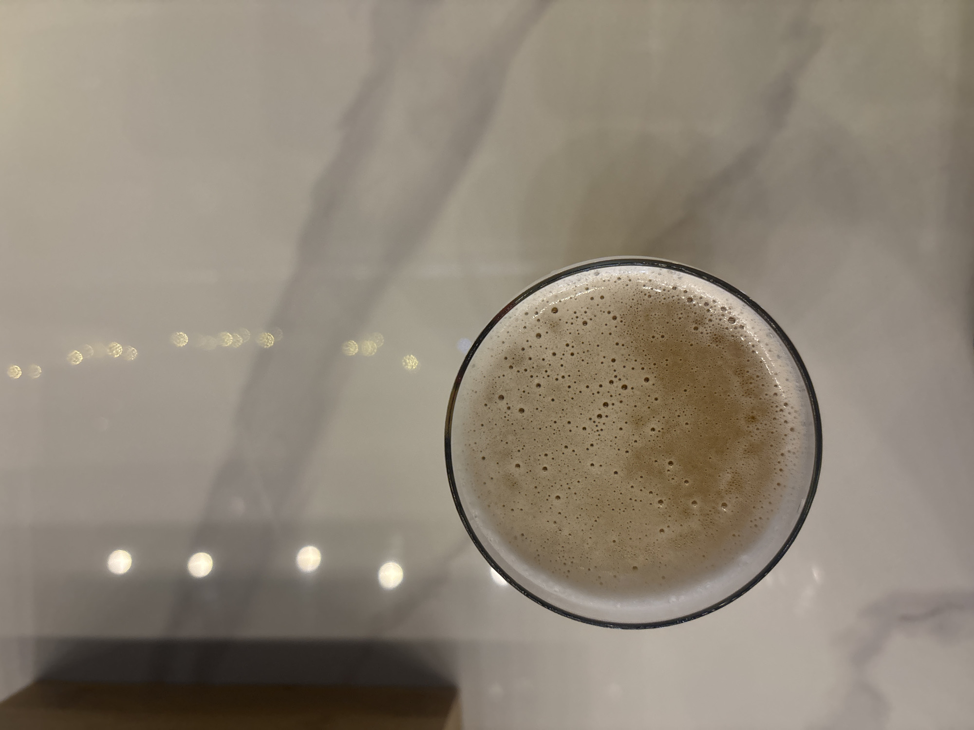 An overhead, medium shot captures a glass of foamy, light brown liquid on a white marble countertop. The glass is positioned slightly left of center and is mostly full, with a thick layer of foam on top. The surface of the foam is speckled with tiny bubbles. The marble countertop features subtle grey veining. In the background, to the left of the glass, a wooden surface is visible, casting a shadow. Above the countertop, several circular bokeh lights are scattered, suggesting an indoor setting with ambient lighting. The scene is static and devoid of any visible people or activity.