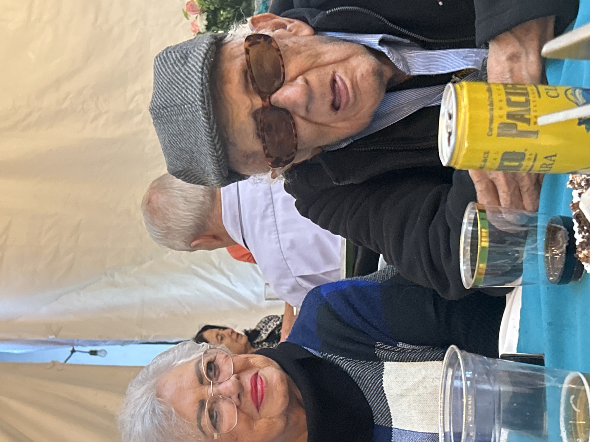 The image shows two elderly individuals seated at a table, presumably in Puebla, Mexico, based on the presence of a can of "Pacífico" beer, a brand commonly associated with Mexico. An elderly woman with white hair, wearing glasses and red lipstick, is in the foreground. In the background, an elderly man wearing a gray hat and sunglasses is visible. A can of "Pacífico" beer and several plastic cups are on the table. Another individual with their back to the camera is wearing a white shirt and an orange shirt beneath. Another person is sitting behind the first woman. The setting is outdoors, under a tent or similar covering.