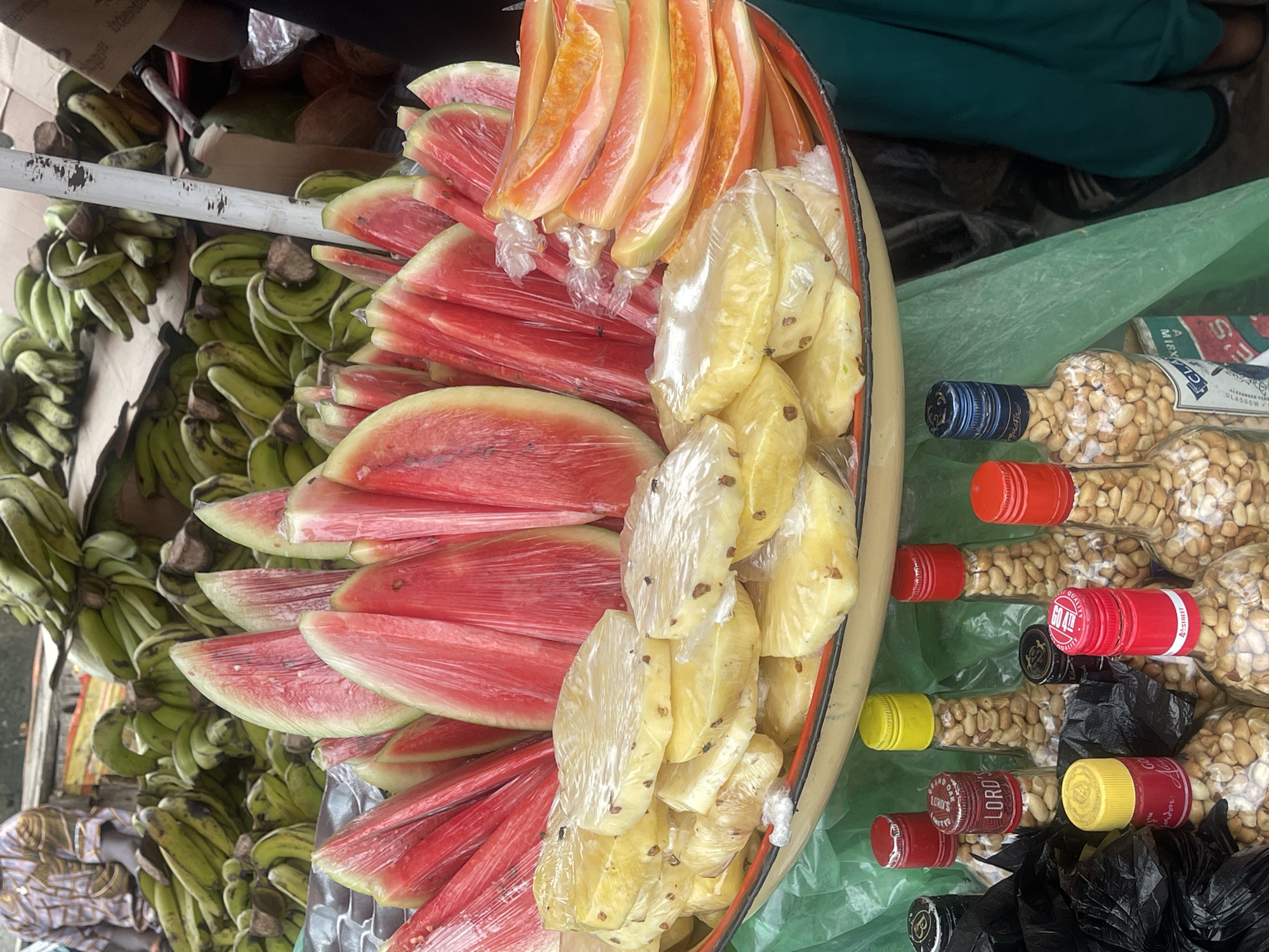 This image shows a street vending setup in Ado, Nigeria. Displayed for sale are a large quantity of green bananas, watermelon slices, pineapple chunks, and papaya slices, all individually wrapped in clear plastic. Glass bottles filled with peanuts and sealed with various colored caps (red, yellow, blue) are also offered. The arrangement of goods suggests a roadside market or informal sales location. The presence of green bananas and wrapped fruit indicates a focus on fresh produce for immediate consumption. The person selling the bananas is wearing a brightly-colored shirt. The environment is urban and somewhat cluttered.