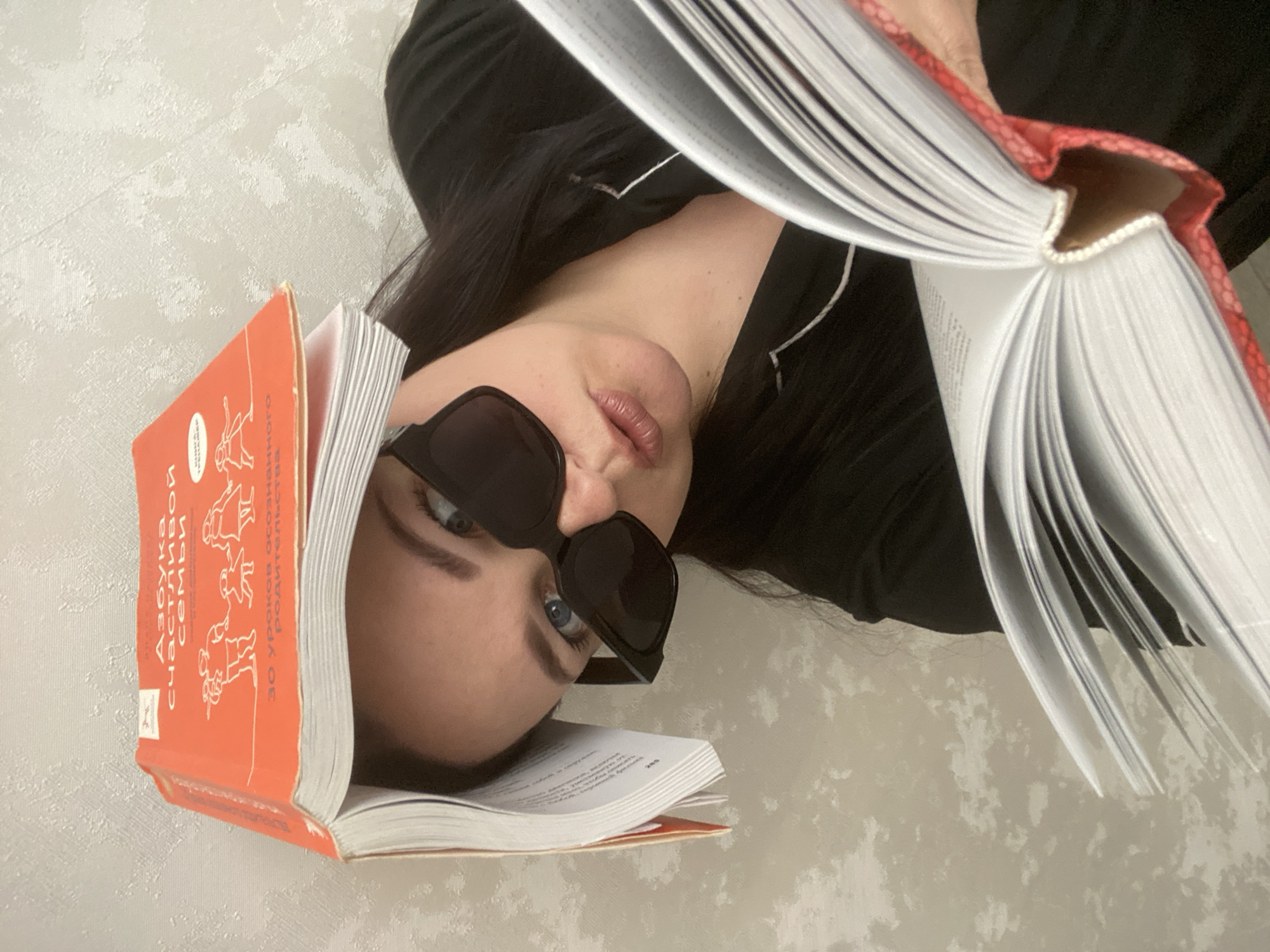 An overhead, close-up photograph captures a person lying down, with the image rotated 90 degrees clockwise. The subject, appearing to be a young woman with long dark hair, is wearing a black top. They have dark sunglasses resting on their nose, revealing striking blue eyes that gaze directly at the camera. Their lips are slightly parted.
Two open books with vibrant red/orange covers are central to the composition. One book rests upon the person's forehead, its orange spine visible. The cover displays Cyrillic text, clearly readable as "АЗБУКА счастливой семьи" (Alphabet of a Happy Family) and "30 уроков осознанного родительства" (30 lessons of conscious parenting). The second book is held open, partially obscuring the lower half of the person's face. This book features a red, textured cover, possibly with a snakeskin pattern on its spine. The fanned white pages of both books are prominent.
The background consists of a light-colored wall with a subtle, textured pattern, suggesting wallpaper or a specific paint finish in an indoor setting. The image conveys a relaxed and casual atmosphere, possibly depicting a moment of reading or a playful pose. No specific time of day or outdoor weather cues are present. The location context for this scene is Sochi, Russia.