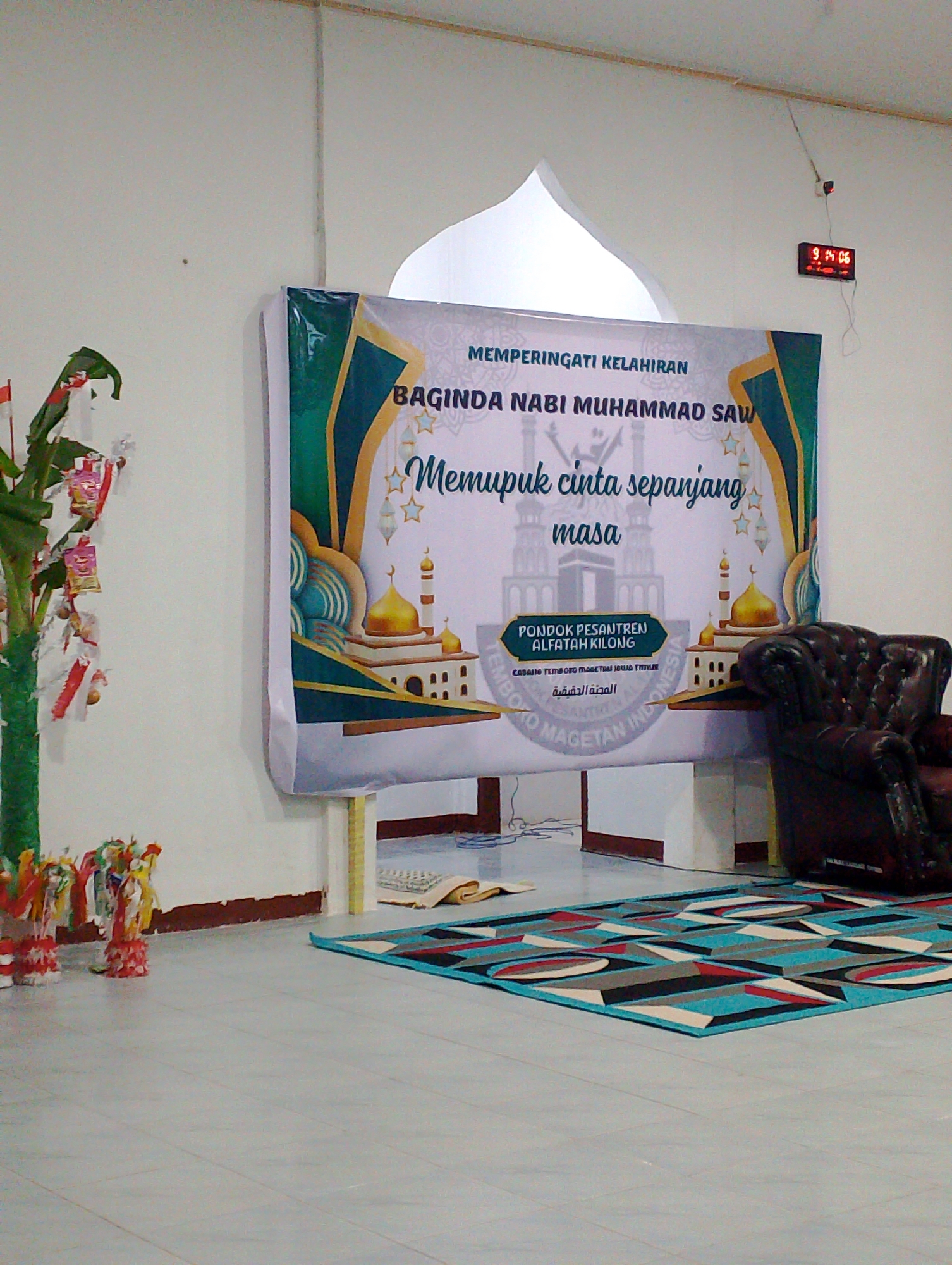 The image shows an indoor setting, likely a community hall or an event space, with a large banner displayed prominently. The banner features text and imagery related to a religious event. To the left of the banner is a green, tree-like decoration adorned with red and white ribbons and small packets. On the right side of the banner, a dark brown leather armchair is positioned. The floor is tiled with a geometric pattern rug in front of the banner and chair. In the background, a white arched structure is visible, possibly a decorative element of the building. A digital clock displaying "9:14:06" is mounted on the wall above and to the right of the banner. The overall scene suggests a celebration or commemoration is taking place. There is no discernible indication of the city of Bobong, Indonesia, within the image itself.