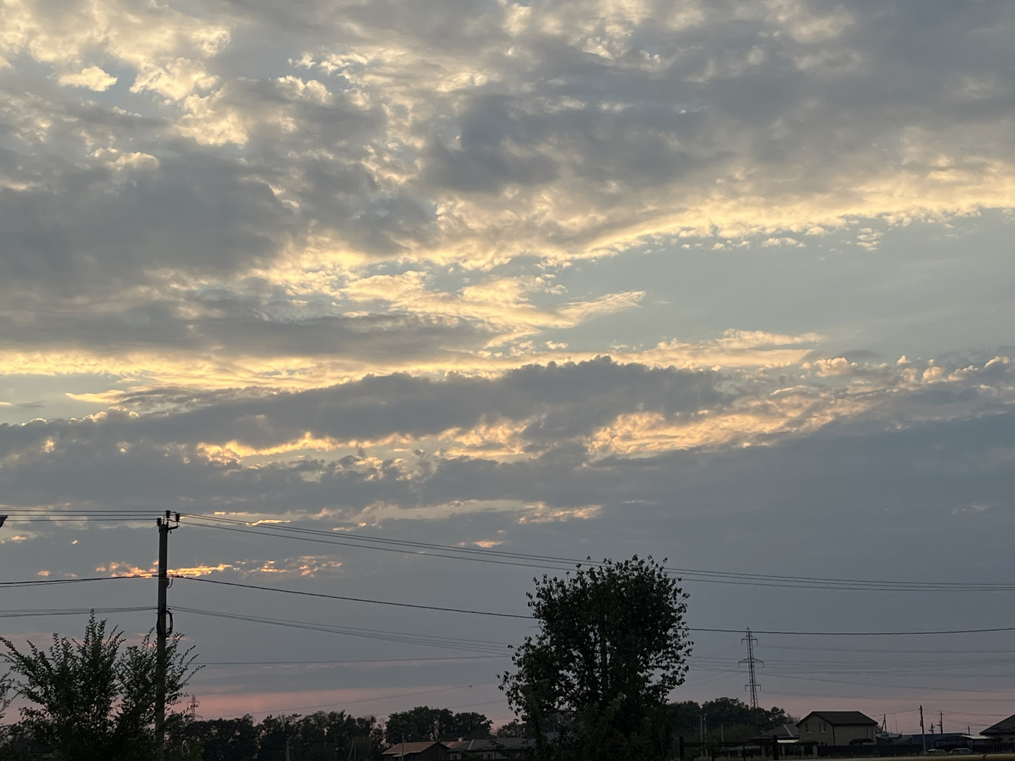 The image depicts a wide, horizontal shot of a sky filled with striated clouds at sunset, casting warm golden and pink hues. In the foreground, a rural landscape unfolds, characterized by silhouetted trees and vegetation. A utility pole with multiple wires stretches across the frame from the left side, partially obscuring the sky. In the lower section of the image, several low-rise buildings are visible, suggesting a residential area. The overall impression is that of a serene, twilight scene in a rural setting, possibly on the outskirts of Taganrog City District, Russia.