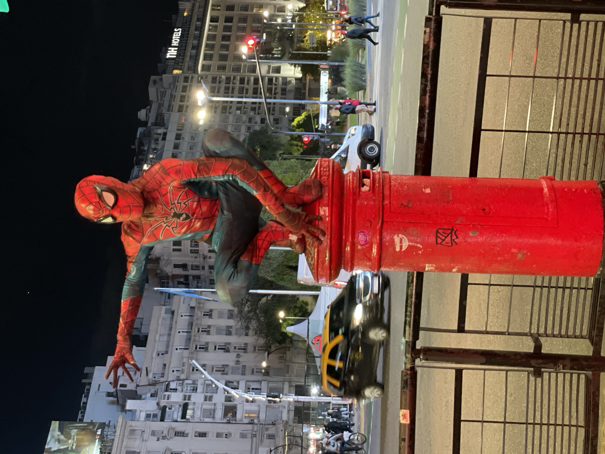 A street performer dressed as Spiderman is perched atop a red cylindrical object, likely a mail collection box. The backdrop consists of city buildings, streetlights, and pedestrian traffic. A black and yellow taxi cab is parked on the street. In the background, "NH HOTELS" is visible on a tall building. Pedestrians are walking in the crosswalk. The image appears to have been taken at night in Buenos Aires, Argentina.