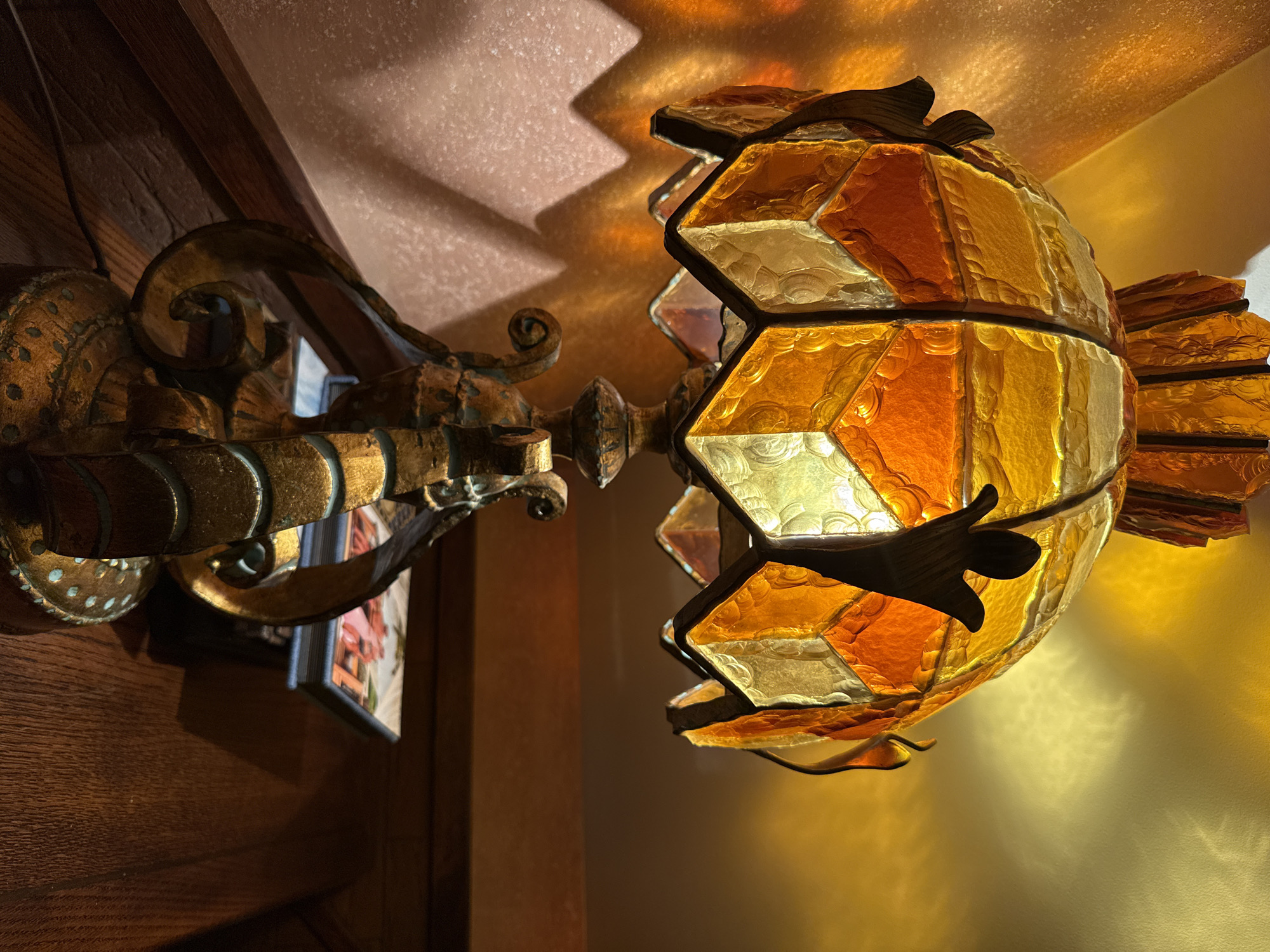The image depicts an ornate, aged sconce with a multifaceted glass shade. The sconce's mounting is gold with decorative scrolling. The shade is composed of amber, orange, and clear glass pieces. The fixture is illuminated, casting jagged shadows on the ceiling above. The sconce is mounted on a wooden wall. A framed picture is visible behind the sconce's arm. The image's location, Lesnoi Gorodok, Russia, is not discernible from the provided visual information.