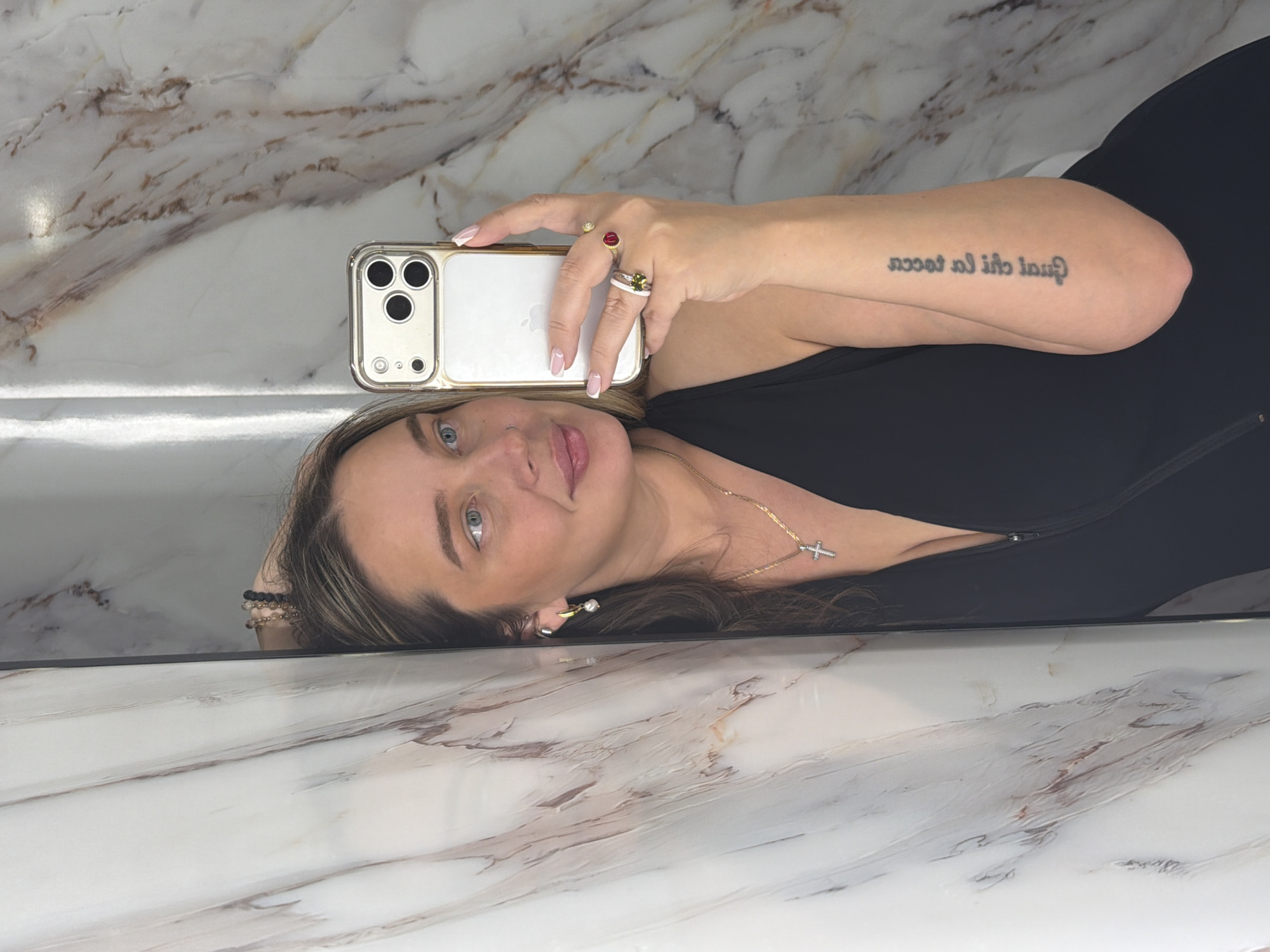 A woman is taking a selfie in a mirrored surface, likely a bathroom. She is lying down, holding a smartphone with a white back and gold trim. She is wearing a black top, a gold necklace with a cross pendant, several rings, and a pearl earring. She has a tattoo on her left arm. The mirrored surface reflects a marble-like background. The location is unknown and not discernible from the image.
