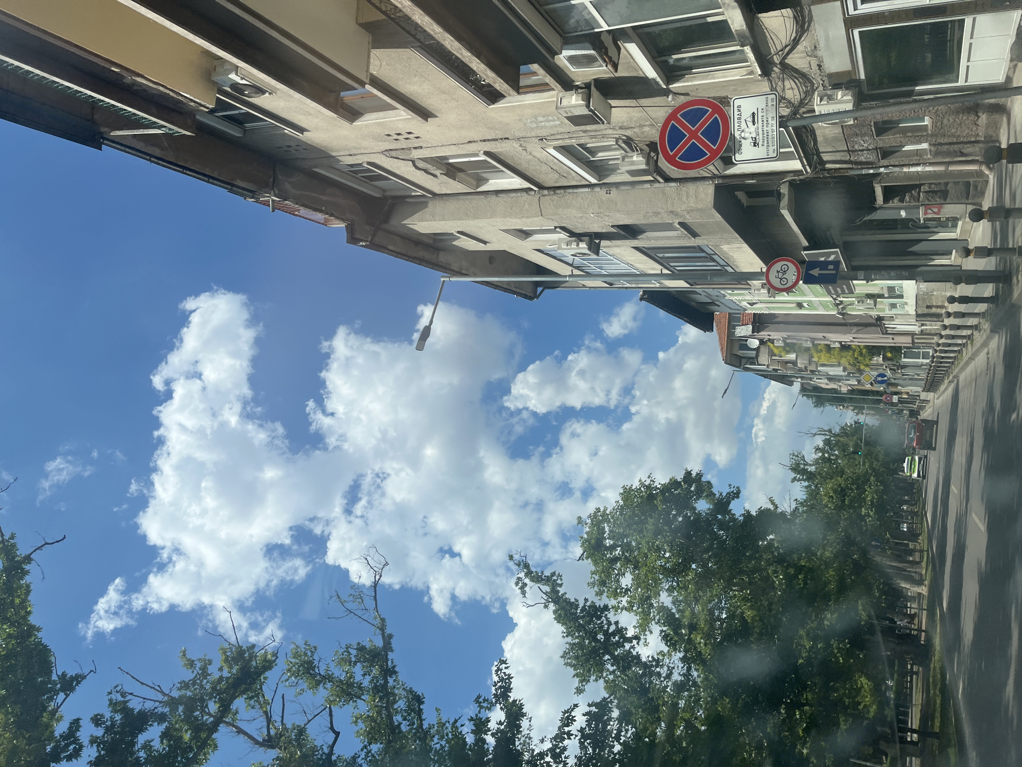 The image depicts a street scene in Plovdiv, Bulgaria, on a sunny day with scattered clouds. The perspective is from a vehicle, looking upwards and towards the right.
On the left side of the frame, an old, multi-story building is visible, with several windows and air conditioning units. Below the building, a bright blue sky with fluffy white clouds dominates the upper portion of the image. Lush green trees with dense foliage line the right side of the street, casting shadows on the asphalt road.
Several traffic signs are present. One prominent sign, a red circle with a blue background and a red cross, indicates "no stopping." Another sign depicts a bicycle, suggesting a bike lane or cycling path. Below these, a white sign with Bulgarian text and a phone number is affixed to a pole. The text on the white sign appears to be: "ОБЩИНА ПЛОВДИВ" (Municipality Plovdiv) and "Нарушавате се от consequências" followed by a phone number and a web address.
Further down the street, other vehicles are seen, including a red car and a green vehicle. The overall impression is a typical urban streetscape on a clear, pleasant day. The time of day appears to be daytime, likely late morning or afternoon, given the bright sunlight and shadows.