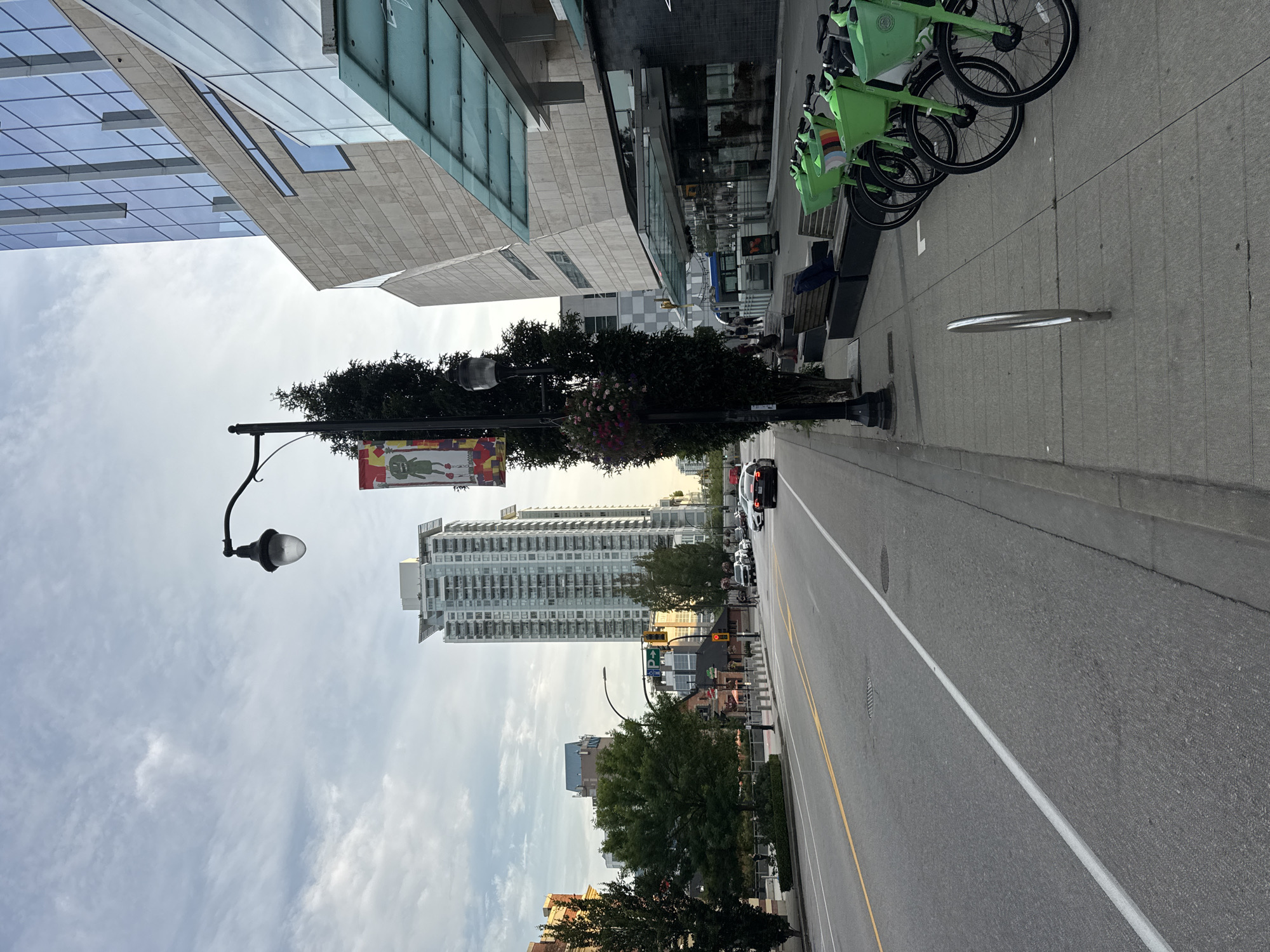 The image depicts a street scene in New Westminster, Canada, on a cloudy day. The perspective is from the sidewalk looking down a wide, paved street with lanes marked by yellow and white lines. On the right side of the street, there is a sidewalk with several bright green rental bicycles parked in a row. A lamppost with hanging flower baskets stands beside the sidewalk, and behind it, a modern building with a stone facade and glass windows rises. Further down the street, a tall, light-colored residential building is visible. Traffic on the street includes a few cars and a dark-colored vehicle in the foreground. Trees are present on the left side of the street, and more buildings and urban infrastructure are seen in the background. The sky is overcast with some wispy clouds.