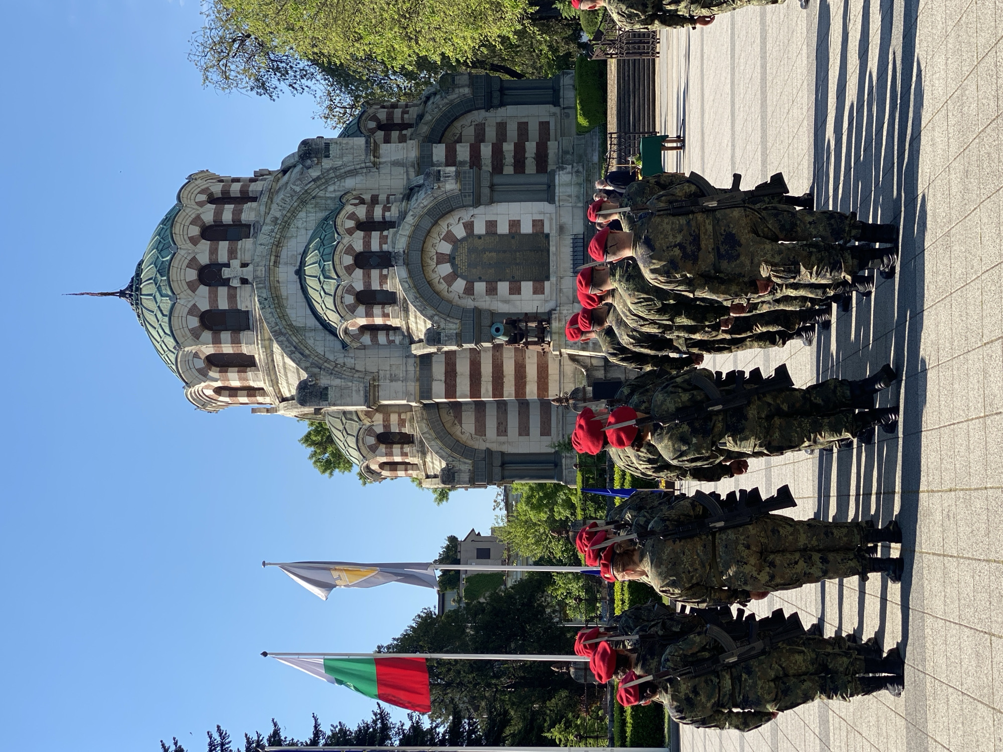 A photo depicts a line of soldiers in camouflage uniforms and red berets standing at attention with rifles, in front of the St George the Victorious Chapel Mausoleum in Pleven, Bulgaria. Flags of Bulgaria are visible. The sky is blue, and the scene is well-lit. The surface appears to be a paved plaza.