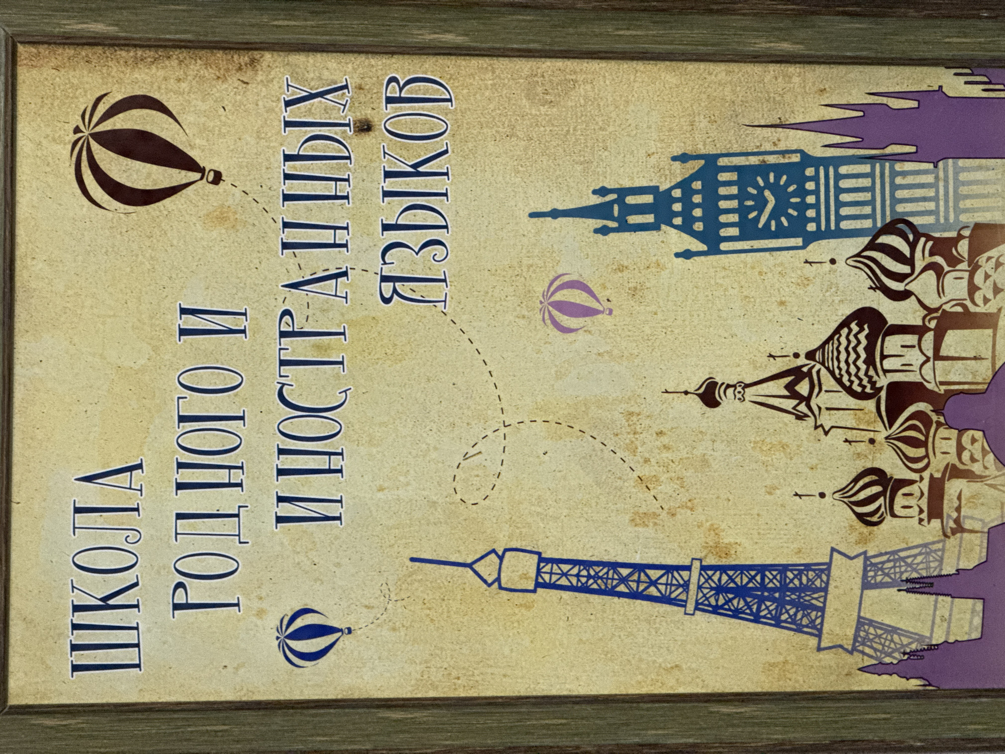 The image is a framed poster for a language school. The text on the poster, written in Cyrillic script, translates to "School of Native and Foreign Languages." The poster's design features several iconic world landmarks, including the Eiffel Tower, Big Ben, and the domes of Saint Basil's Cathedral in Moscow. These landmarks are rendered as stylized illustrations. Hot air balloons are also scattered throughout the design, connected by a dotted line. The color palette consists of muted tones, with blue, brown, and purple accents. The background has a textured appearance, suggesting an aged or vintage aesthetic. The frame surrounding the poster is made of wood, with a green-tinted finish. There is no indication of the location of the city of Votkinsk, Russia in the image.
