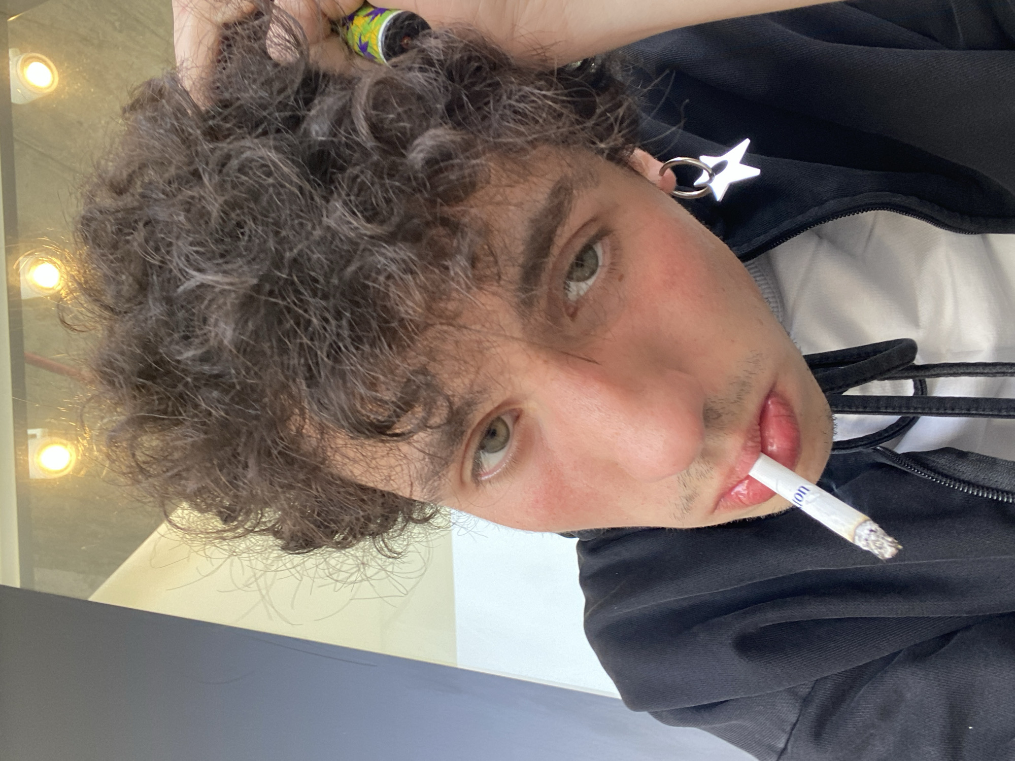A close-up, rotated portrait shows a fair-skinned person with curly brown hair, hazel eyes, and a cigarette held in their mouth. They are wearing a black zip-up jacket over a white shirt. An earring in the shape of a star is visible. The image is taken indoors, with reflections of lights visible in the background. The location in Tel-Aviv, Israel is not discernible from the image.