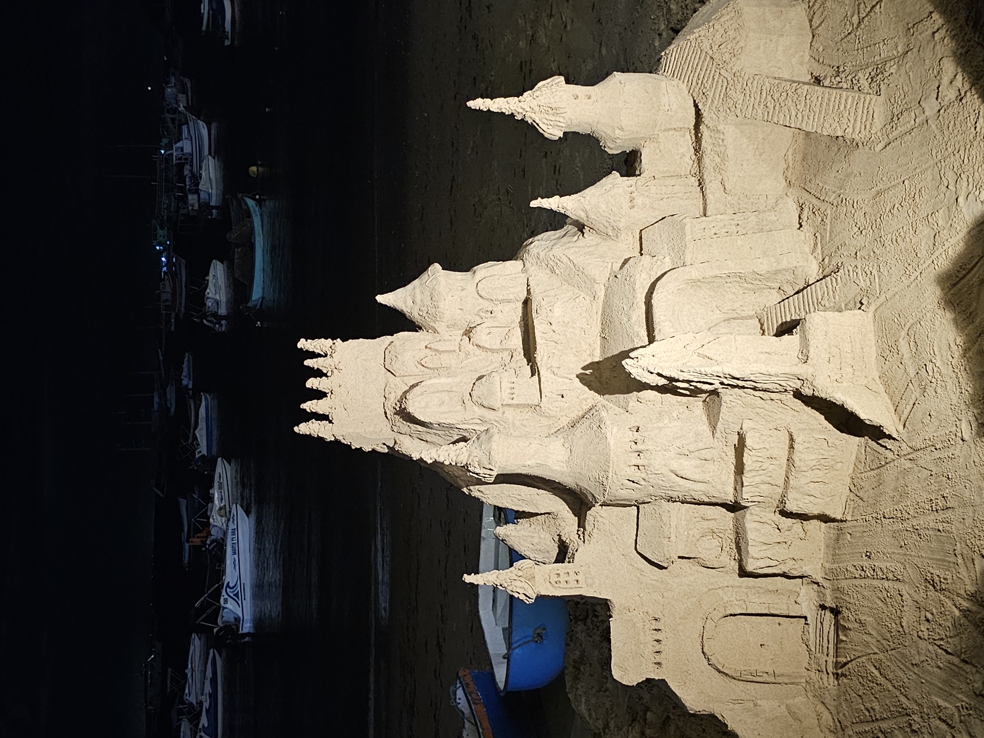A sandcastle stands prominently on a beach at night in Armação Dos Búzios, Brazil. The castle is elaborate with spires, arches, and intricate textures, illuminated by artificial light. Adjacent to the beach is a collection of boats docked along the shoreline. The scene is mostly dark, highlighting the sandcastle as the main subject.