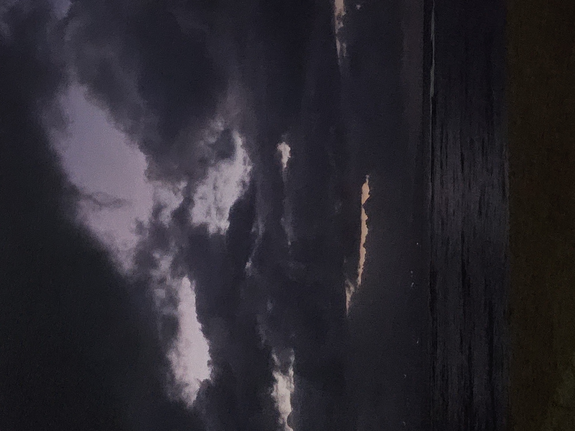 The image shows a dark and cloudy scene, possibly at twilight or night. The left side of the image is dominated by dark, thick clouds with hints of lighter areas suggesting breaks in the cloud cover. The right side appears to show a body of water, with a textured surface that may be due to waves or reflections. There is a dark, solid area on the far right, possibly land or another object obstructing the view. The photo is of the sky and an outdoor area. There is no visible evidence to establish location in the city of Denpasar, Indonesia.