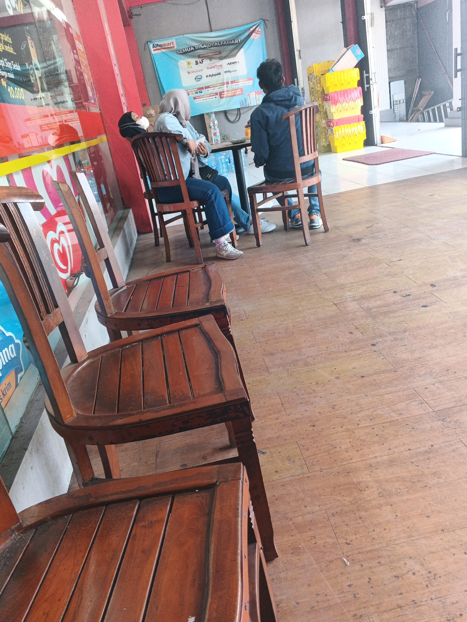 Two individuals, a male and a female, are seated on wooden chairs in front of a storefront that appears to be a minimarket in Genuk Barat, Indonesia. The female, wearing a light blue hijab and denim jeans, is seated on a wooden chair facing away from the camera. She has a white mask covering her nose and mouth. She is looking to her left, down at the floor. The male, wearing a dark blue hooded jacket and blue jeans, is seated on another wooden chair to the right and slightly behind the female. He is also facing away from the camera, looking towards a table between them. Several other empty wooden chairs are visible in the foreground, arranged in a row. Behind the individuals, a blue banner is visible on the wall, displaying the Alfamart logo and various payment options. Yellow and pink plastic containers are stacked near the entrance of the store to the right. The floor is made of wooden planks. The overall scene depicts a casual waiting area outside a commercial establishment.