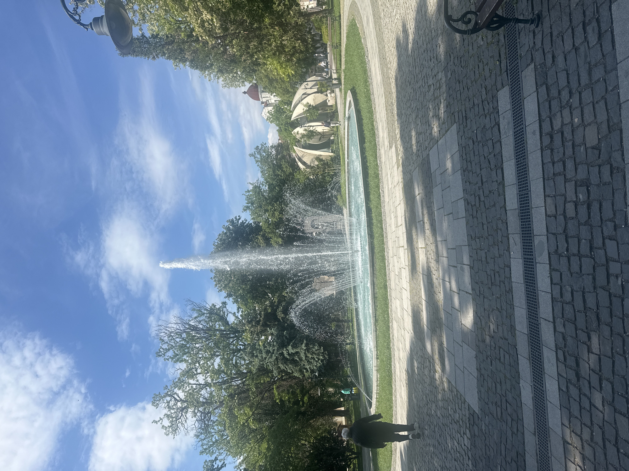 The image depicts a scene in a park in Plovdiv, Bulgaria. A large fountain is the central subject, with a tall jet of water spraying upwards. The fountain is surrounded by green grass and trees. Paved walkways border the fountain. A person is walking along the walkway on the lower right side of the image. In the background, there are buildings with unique architectural designs, including structures with domed roofs. The sky is blue with scattered white clouds. A lamppost is visible at the upper left corner of the image. A bench is partially visible at the upper right corner of the image.