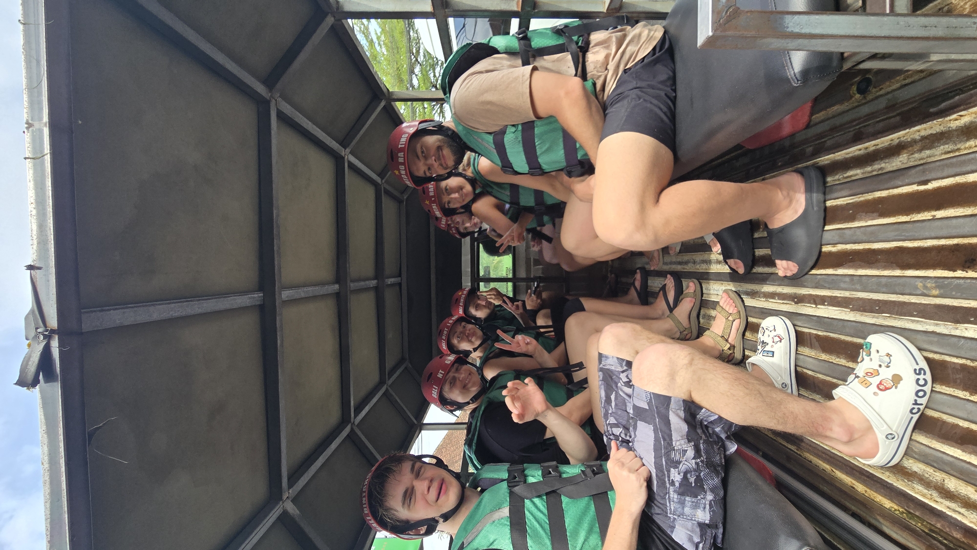 A group of approximately seven individuals is depicted inside what appears to be an open-sided transport vehicle, likely a truck or van. The image is oriented sideways, but if viewed upright, the subjects are seated on long benches facing forward, with their backs against the side of the vehicle. All individuals are wearing green and black life vests and red helmets, suggesting preparation for or return from an adventure activity, such as rafting or kayaking, in the Ubud District, Indonesia.
Most subjects are smiling, and several are making peace signs with their hands, indicating a positive and relaxed mood. Their attire includes casual shorts and t-shirts. The vehicle's interior features a sturdy metal frame structure supporting a ceiling, and a corrugated metal floor. Footwear visible includes various flip-flops, sandals, black slides, and a pair of white Crocs adorned with colorful charms.
Visible text on the red helmets includes "GAYANG RAFTING" and "BALI RIVER RAFTING," confirming the nature of the activity. Another helmet partially shows "BINTAN." The Crocs brand name is also clearly visible on one shoe. The bright lighting suggests it is daytime with clear weather. The vehicle's open design allows glimpses of greenery and blue sky outside, characteristic of an outdoor setting.
