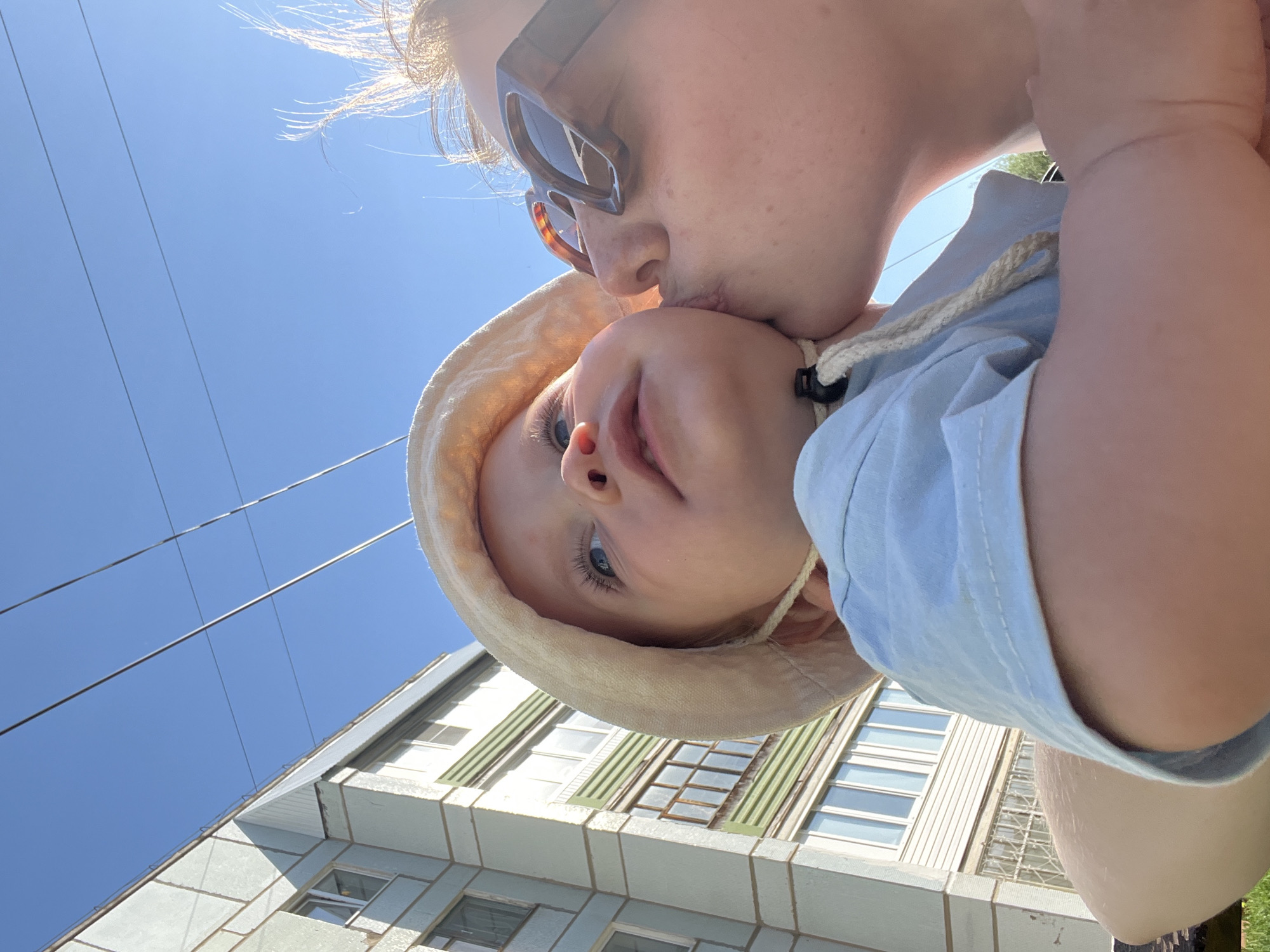 A woman wearing sunglasses kisses a baby wearing a sun hat. They are positioned in front of a building with multiple windows and green shutters. Above them is a clear blue sky with power lines. The location appears to be an urban area, possibly in the city of Chaikovsky, Russia.