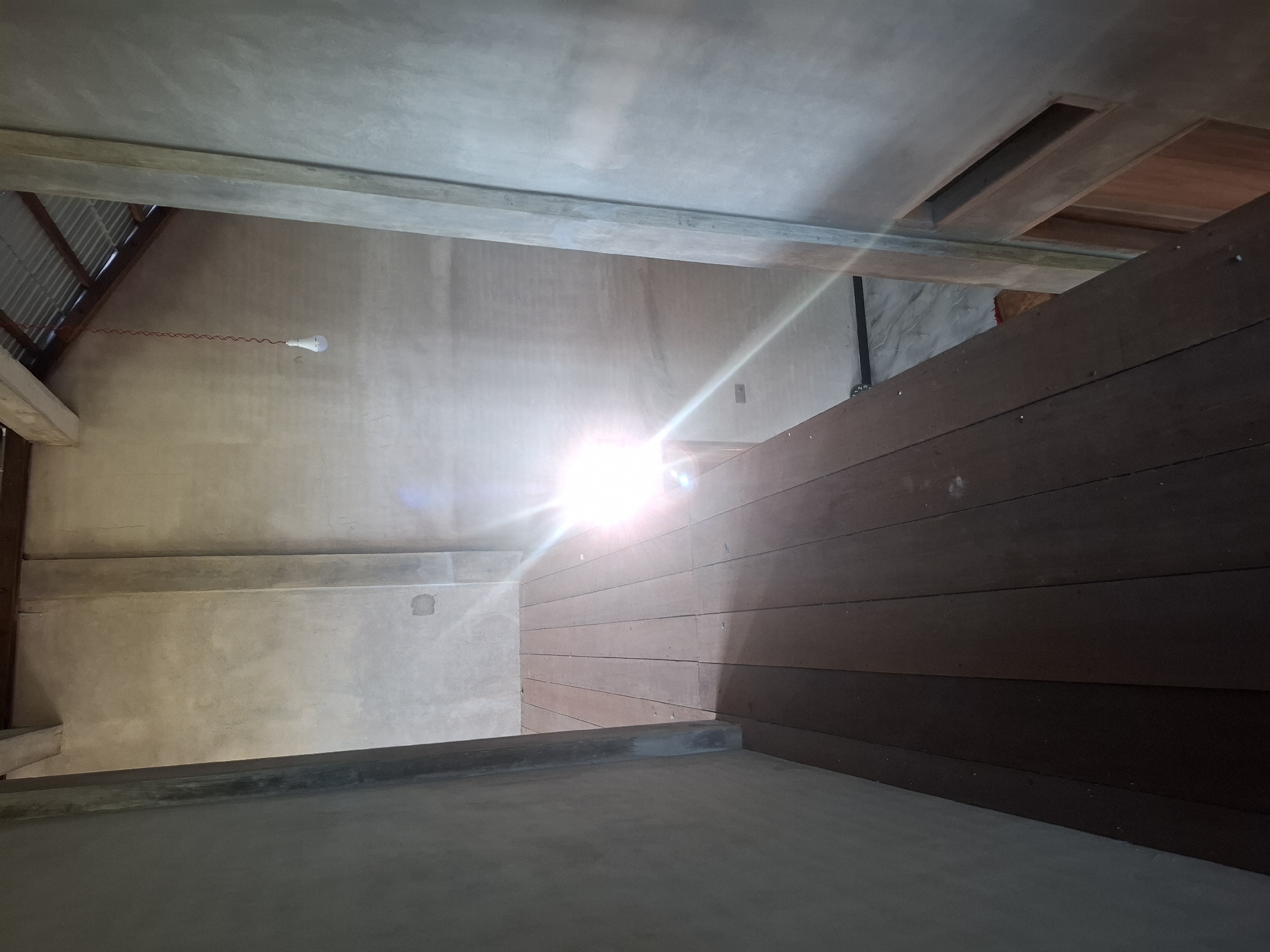The image shows the interior of a partially constructed building, likely a house, in Gronggrong, Indonesia. The scene is viewed from a wooden walkway or balcony, looking down into a larger space. The walls are unpainted concrete, and a corrugated metal roof is visible in the upper right. A bare light bulb hangs from a red wire, illuminating the space. The wooden floorboards of the walkway are visible in the foreground, with a bright sun glare obscuring details in the middle ground. Below the walkway, a marble-tiled floor and a wooden door are partially visible. The overall impression is one of a rustic, unfinished structure.