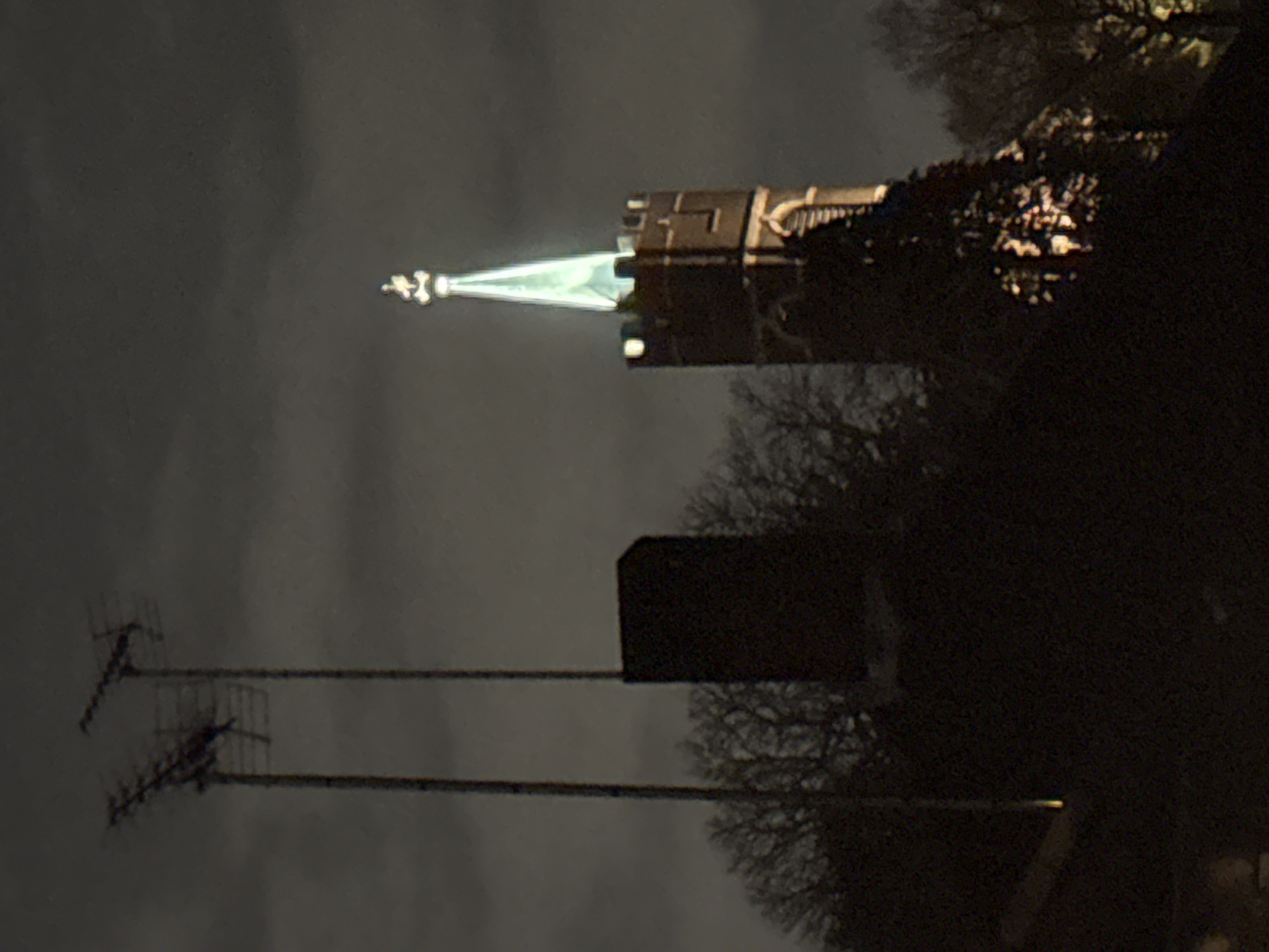 The image depicts a nighttime scene in Putney, London, United Kingdom, specifically featuring the upper section of St Mary's Church, Putney (SW15 1SN). The church's brick or stone tower is visible, topped with battlements, and supporting a tall, slender octagonal spire that is brightly illuminated with a greenish-white light. A finial crowns the spire.
To the left of the church, two tall, dark poles rise from beyond the lower frame, each supporting a multi-element directional antenna, likely for television reception. A dark, rectangular building or structure is partially visible between the antennae and the church tower. Bare deciduous trees, indicating a winter setting, are silhouetted against the dark, overcast sky, surrounding the structures and extending across the lower portion of the frame. Artificial light illuminates the lower section of the church tower and parts of the trees to the right with an orange-yellow hue. A small, bright yellow light source is present at the bottom left edge of the image. The sky is dark grey with visible cloud cover. No individuals or vehicles are present.
