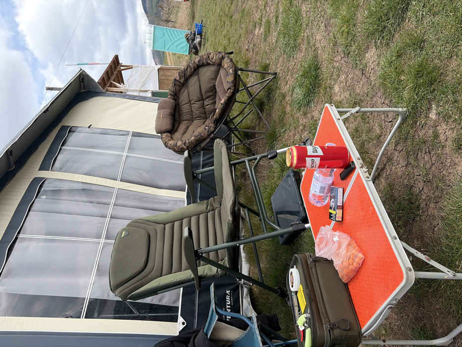 This image captures an outdoor scene within the specified city of Cherna Gora, Bulgaria, under an overcast sky. In the foreground, an unoccupied green, padded camping chair is positioned to the left of a small, orange-topped, silver-legged folding table. On the table, from left to right, are an open clear plastic bag containing multiple orange, spherical food items; a blue and white cigarette pack labeled "Marlboro Gold"; a black utility knife or lighter; a clear plastic water bottle lying on its side; and a red beverage can with white lettering, consistent with a beer brand. An olive green soft-sided utility bag, containing a white spool and other small items, rests on the ground to the left of the table. To the right of the green chair, an unoccupied brown, camouflaged, padded, rounded camping chair is visible. Behind these items, a large, light beige canvas tent or caravan awning with clear plastic windows forms the backdrop. In the mid-ground, to the right, a partially covered wooden structure and a light blue portable toilet are present. Distant rolling hills are visible under the cloudy sky, and a Bulgarian flag is faintly discernible on a pole behind the wooden structure. No individuals are visible in the scene.